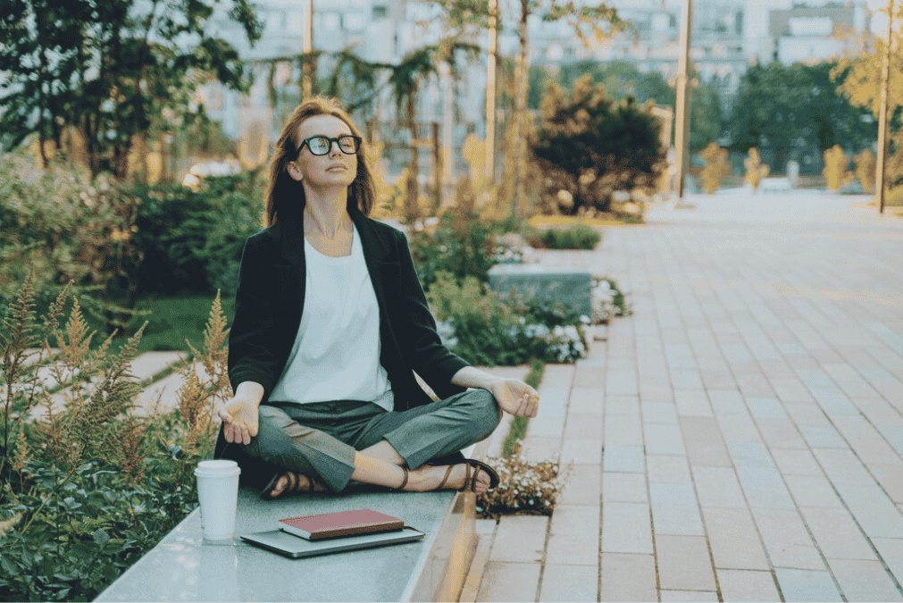 A woman sitting cross-legged on a park bench in a meditative pose, with a coffee cup and a notebook nearby, in an urban park with trees and pathway.