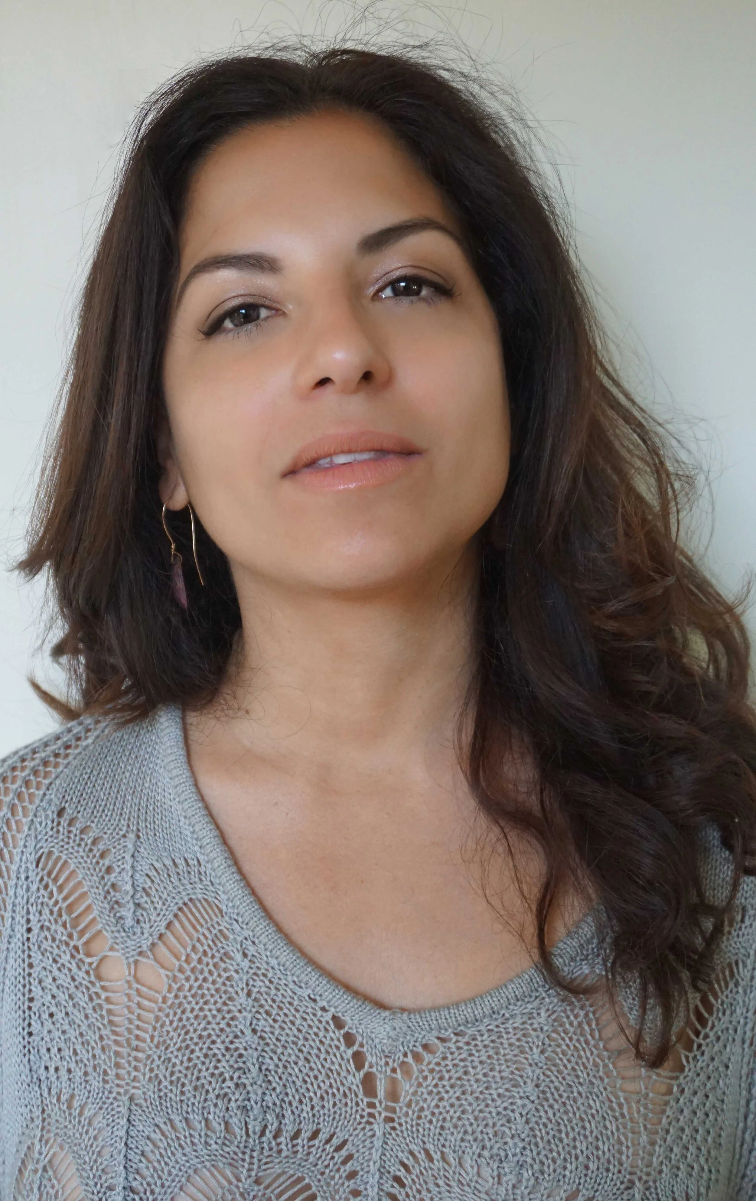Close-up of a woman with dark, wavy hair, wearing a gray knitted top and a pink earring.