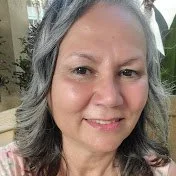 Close-up of a smiling woman with gray hair, outdoors with green foliage in the background.