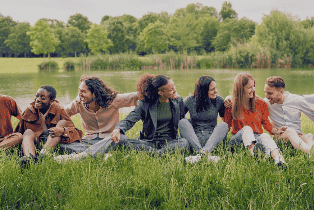 Group of diverse friends sitting on grass by a lake, smiling, enjoying a sunny day.
