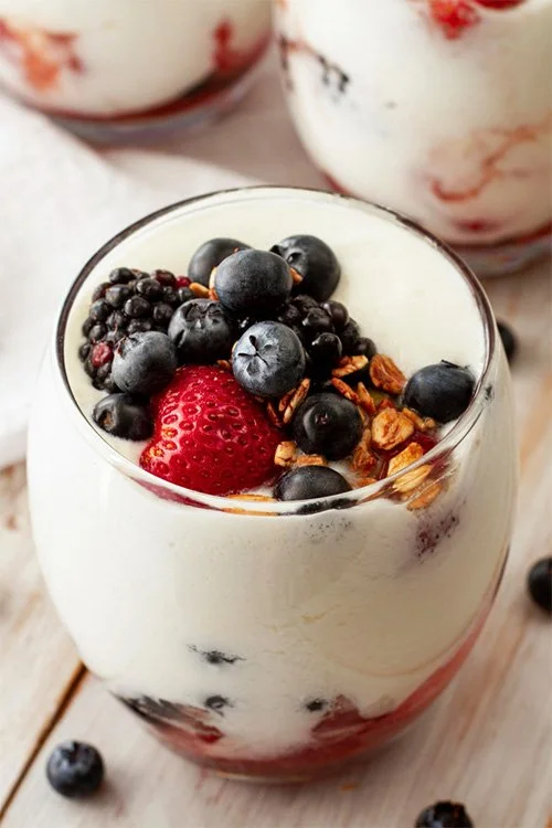 Glass of yogurt topped with blueberries, a strawberry, granola, and other mixed berries, with additional berries around the glass on a wooden surface.