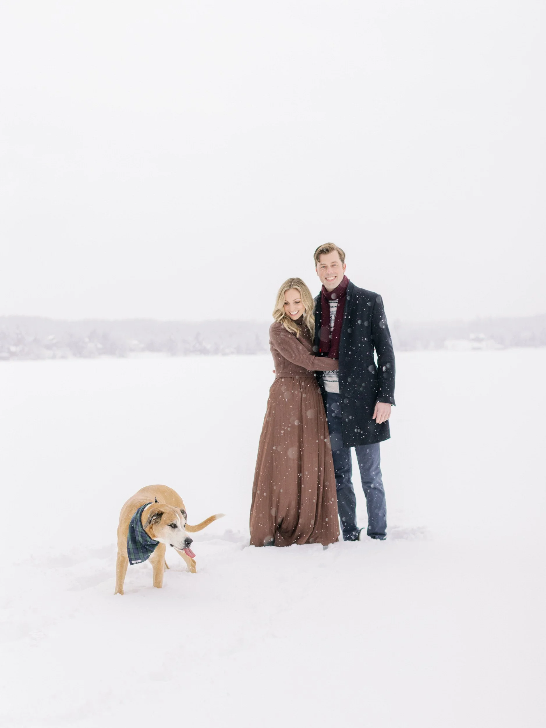 A couple standing in the snow with a dog in front of them