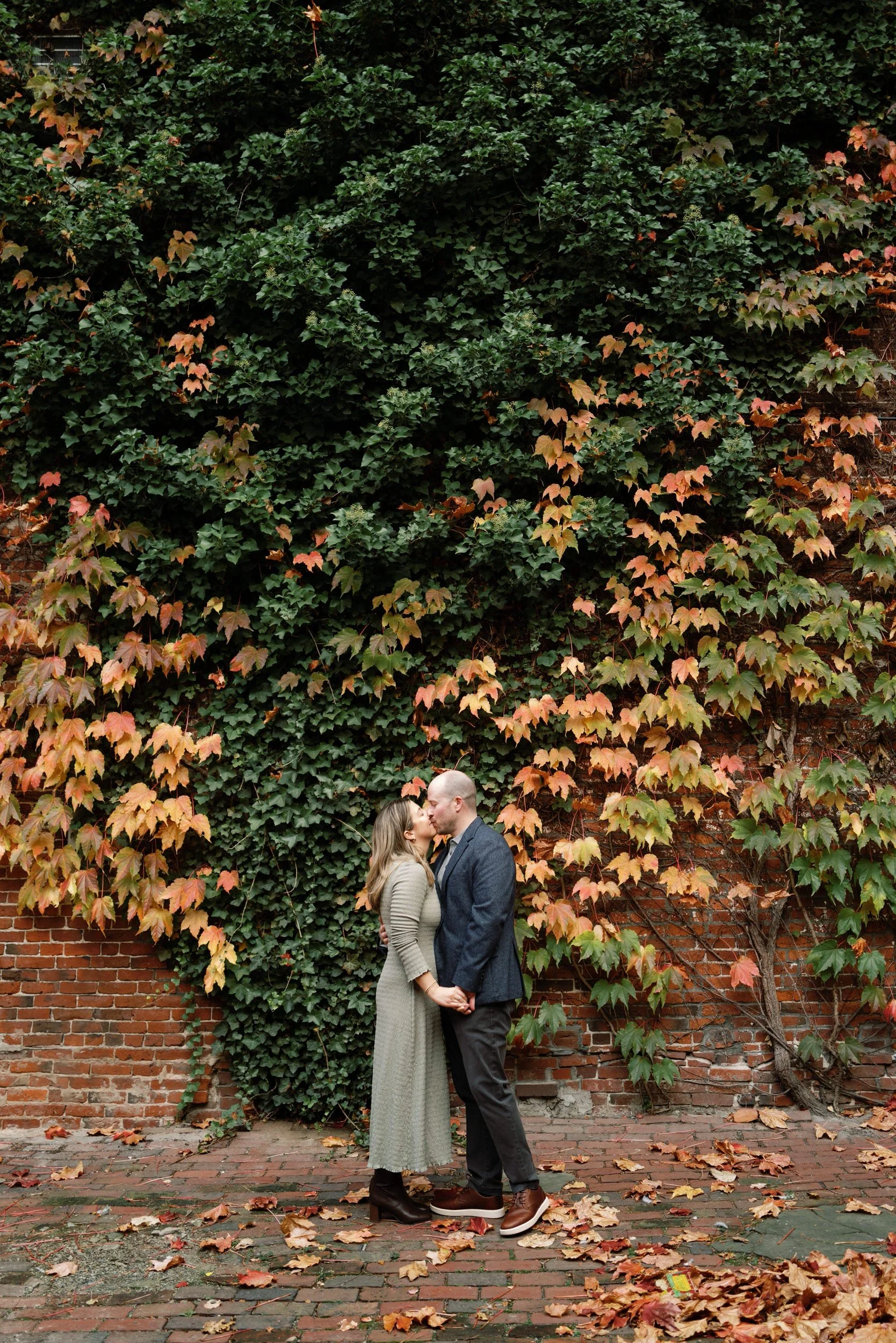 A couple kissing in front of an ivy covered brick wall 