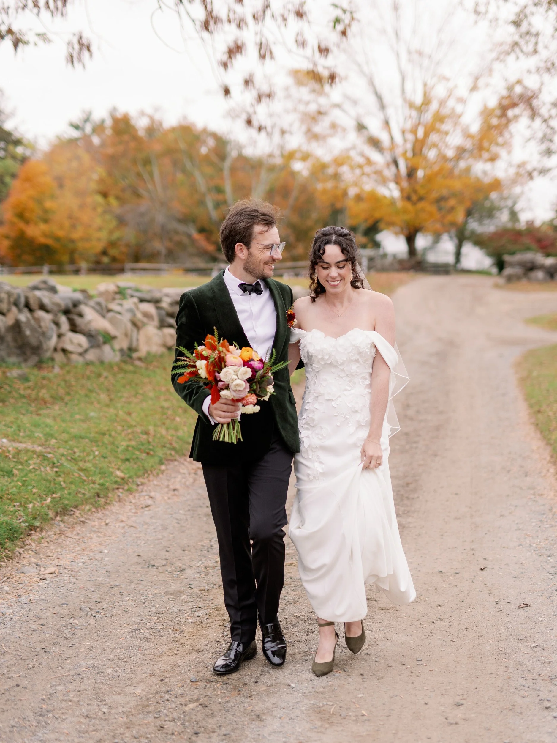 A newlywed couple walking along a gravel road as one holds the other's bouquet of flowers 