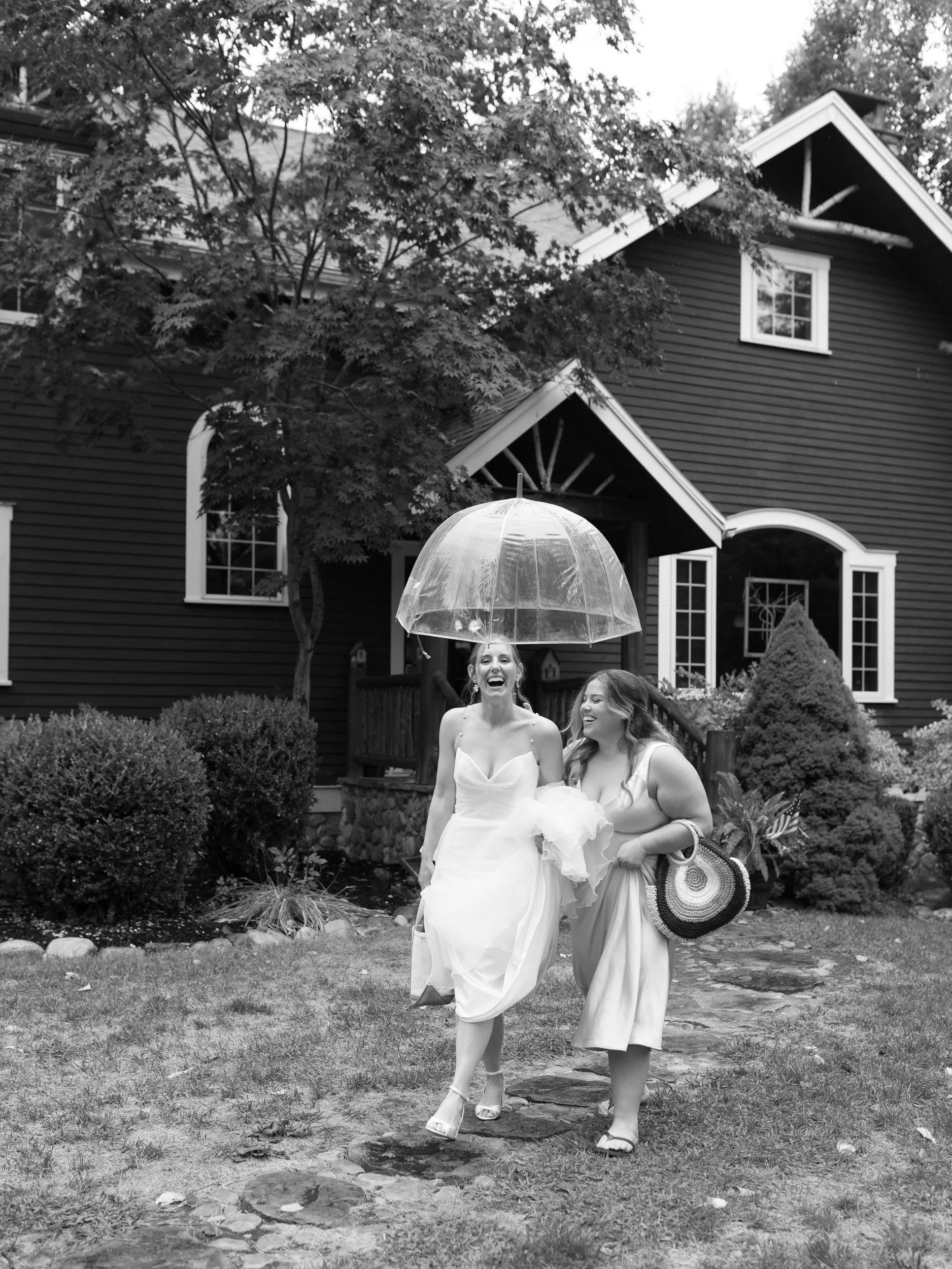 A person in a wedding dress walking across a lawn with a wedding guest under an umbrella 
