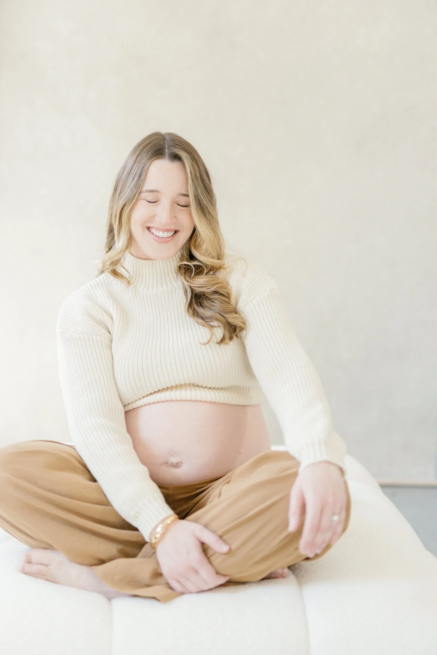 A pregnant person sitting on a white couch smiling 
