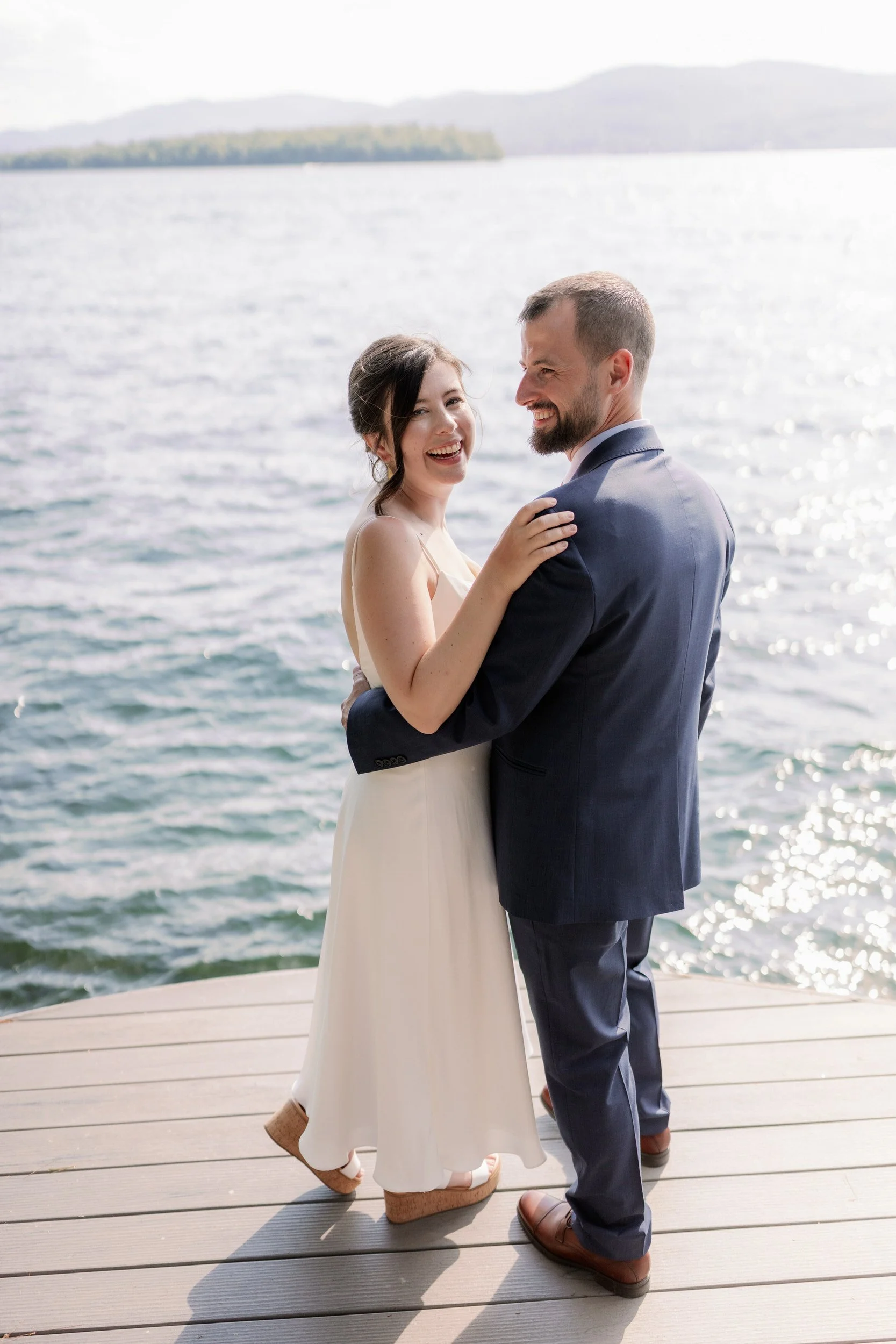 A newlywed couple with their arms around each other smiling on a dock 