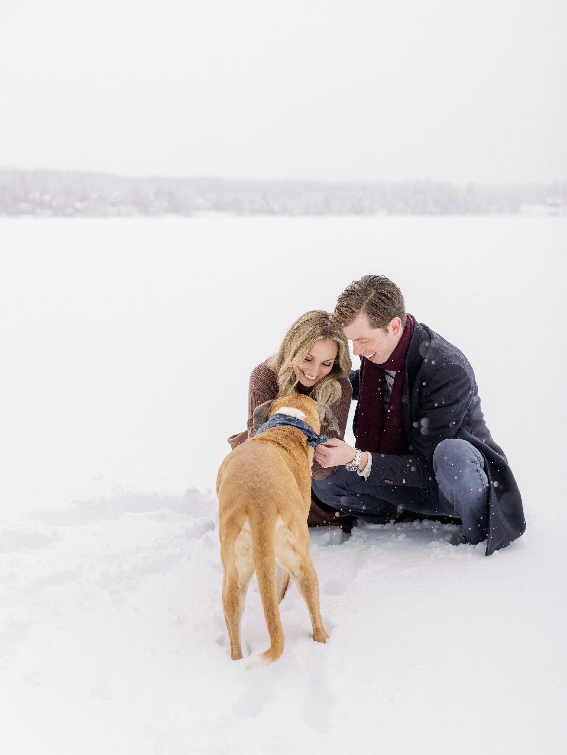 A couple sitting in the snow petting a dog 
