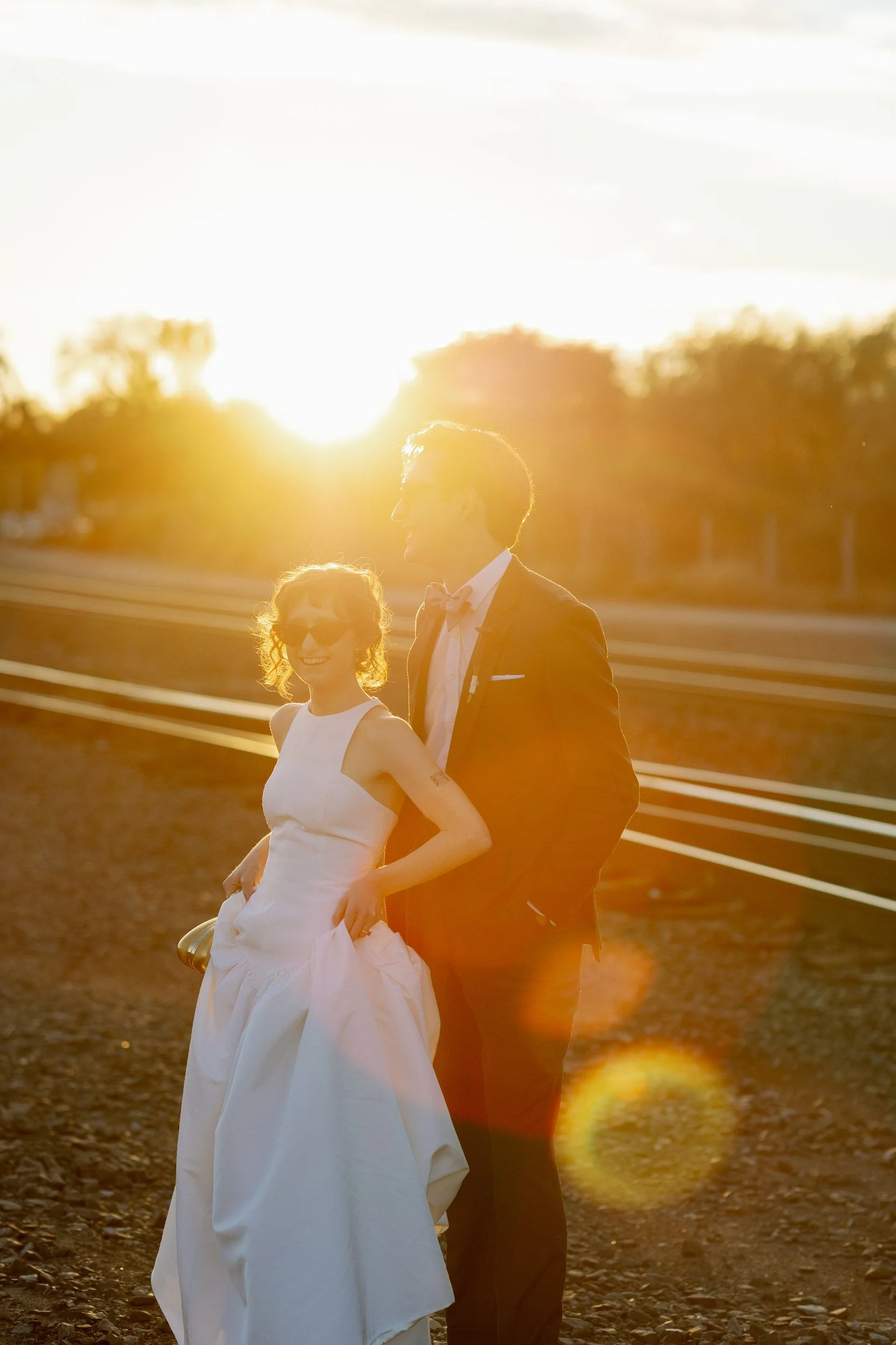  Newlyweds standing one in front of the other smiling in sunglasses at sunset 