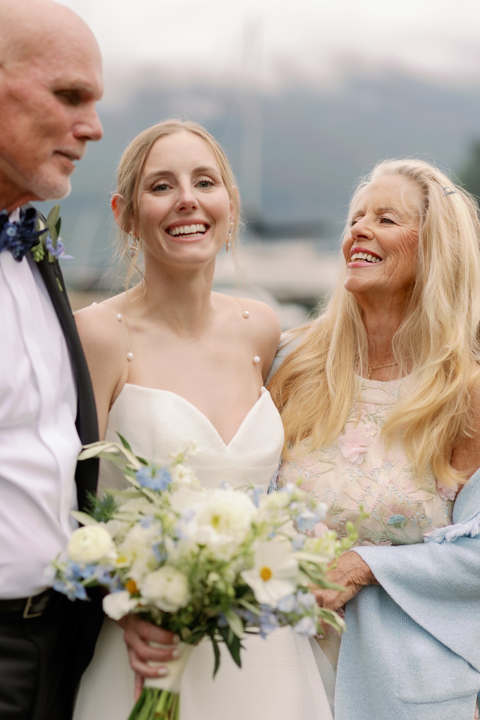 A newlywed in a wedding dress smiling with their parents on their side of them 