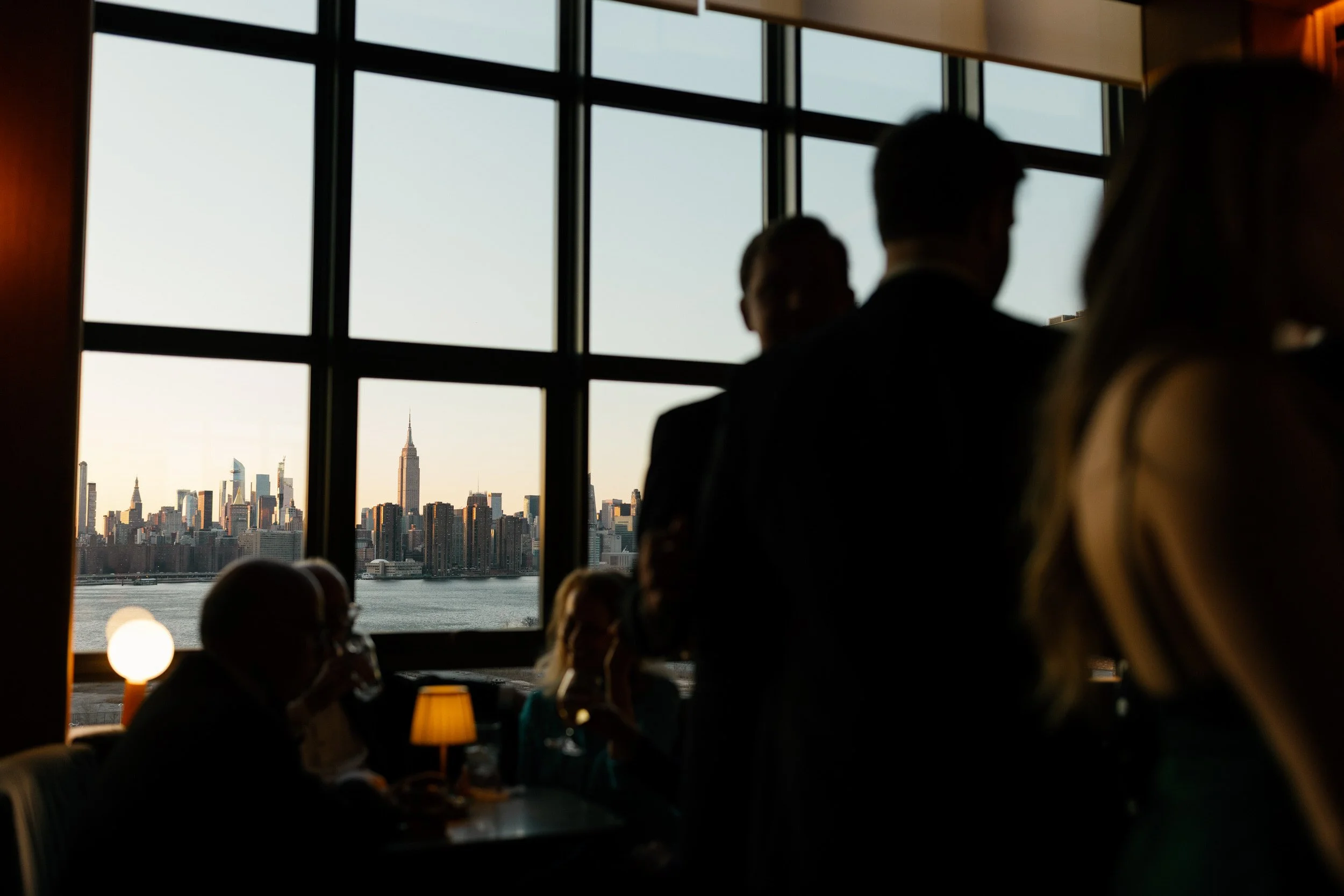 Wedding guests mingling at a wedding reception with the NYC skyline seen through the window in the distance 