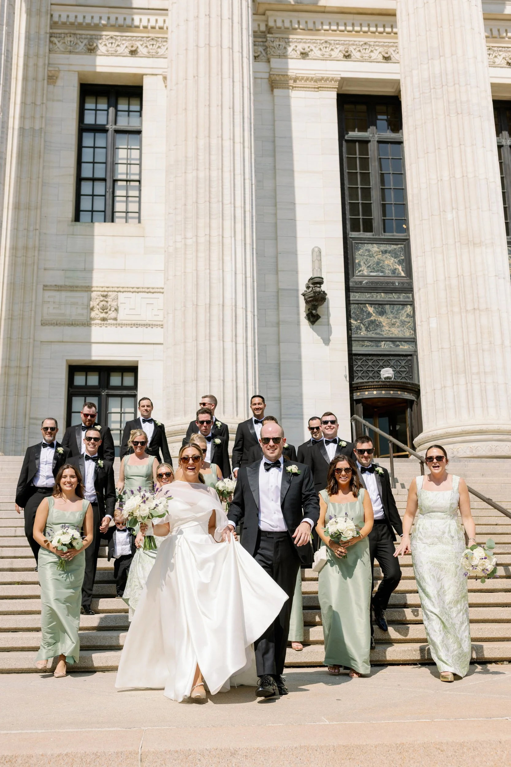 Newlyweds walking down a staircase in front of a building with their wedding parties behind them