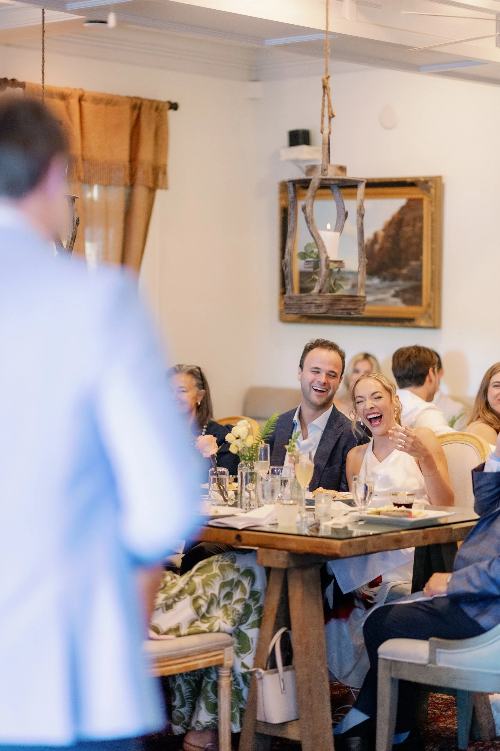 Newlyweds laughing as someone gives a speech at their reception 