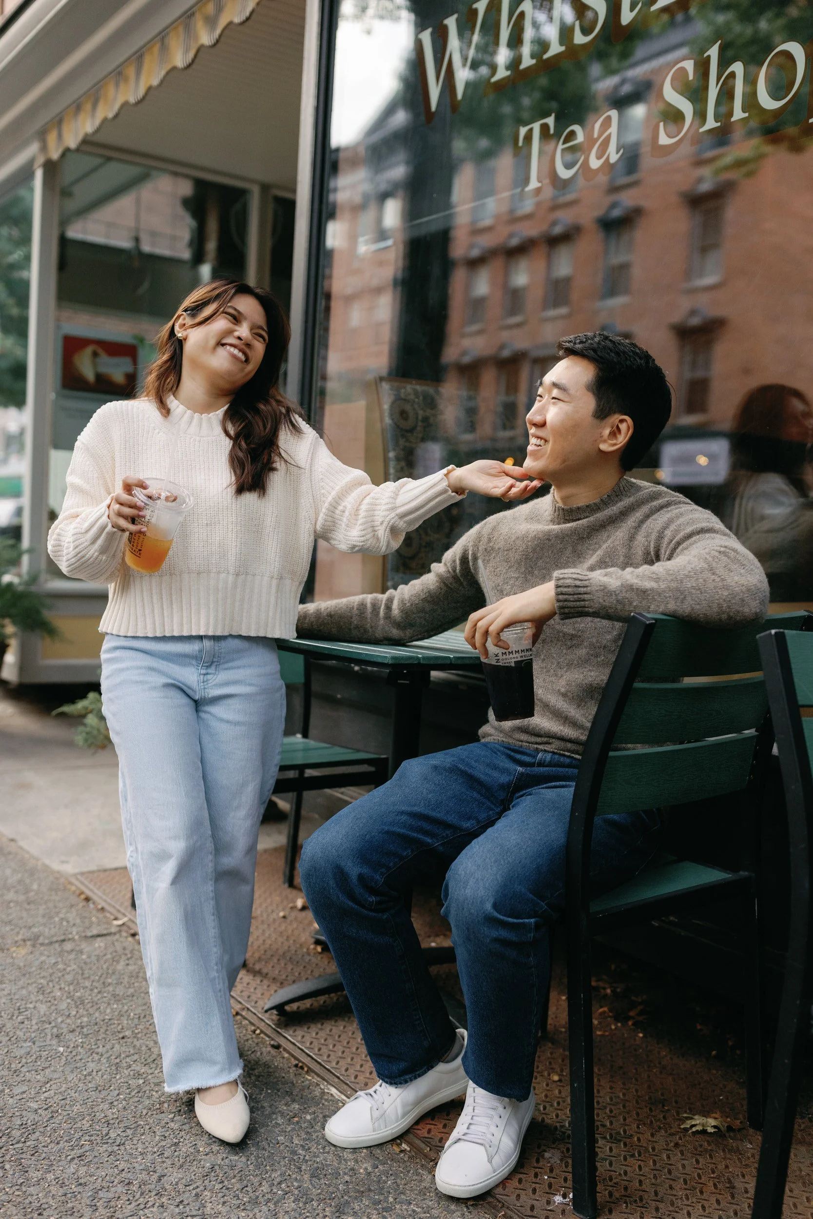 A person sitting at an outdoor cafe table with their partner standing next to them laughing with them