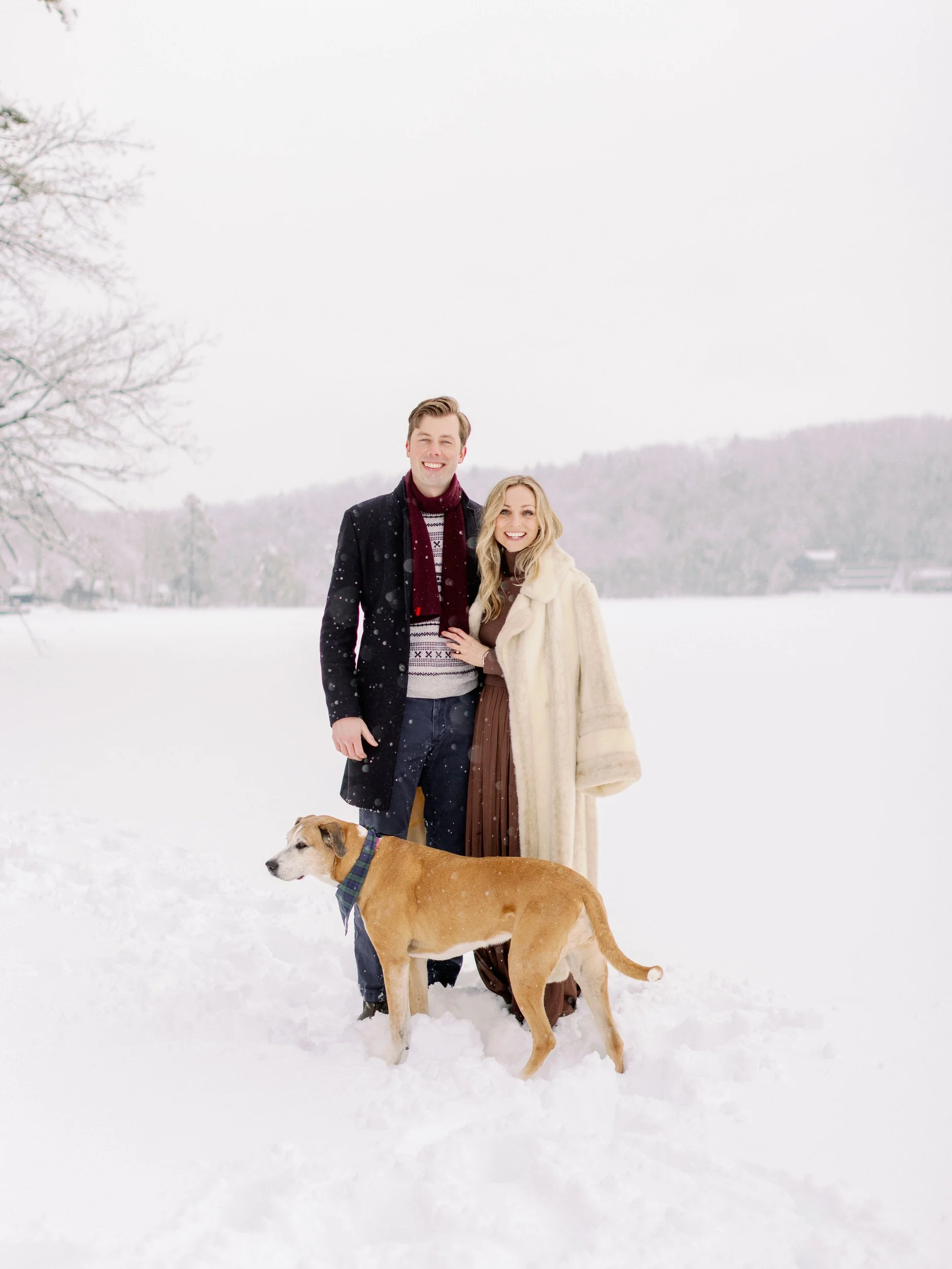 A couple standing in the snow together smiling with their dog in front of them 