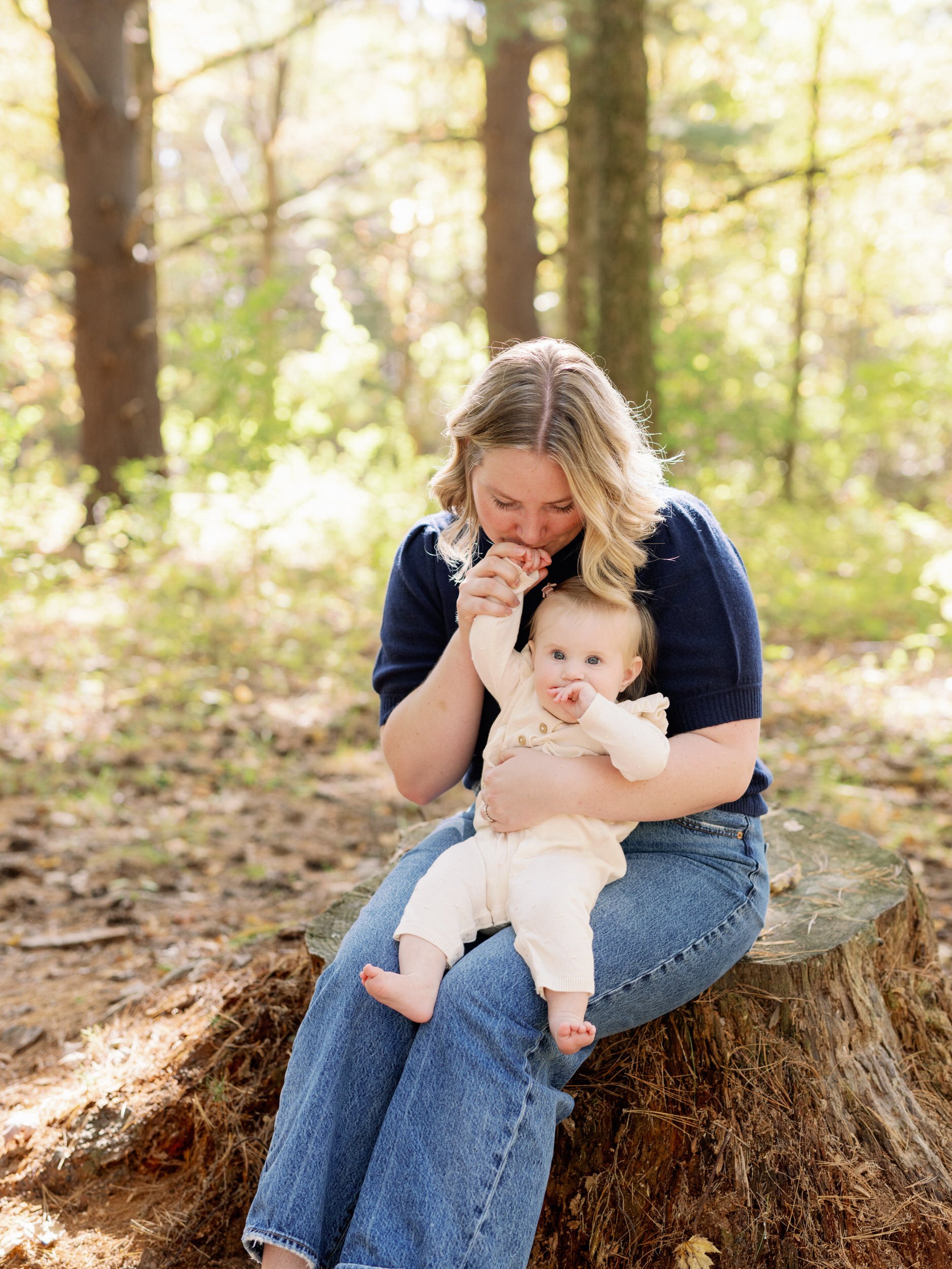 A parent sitting on a tree stump with a newborn in their lap as they kiss their hand 