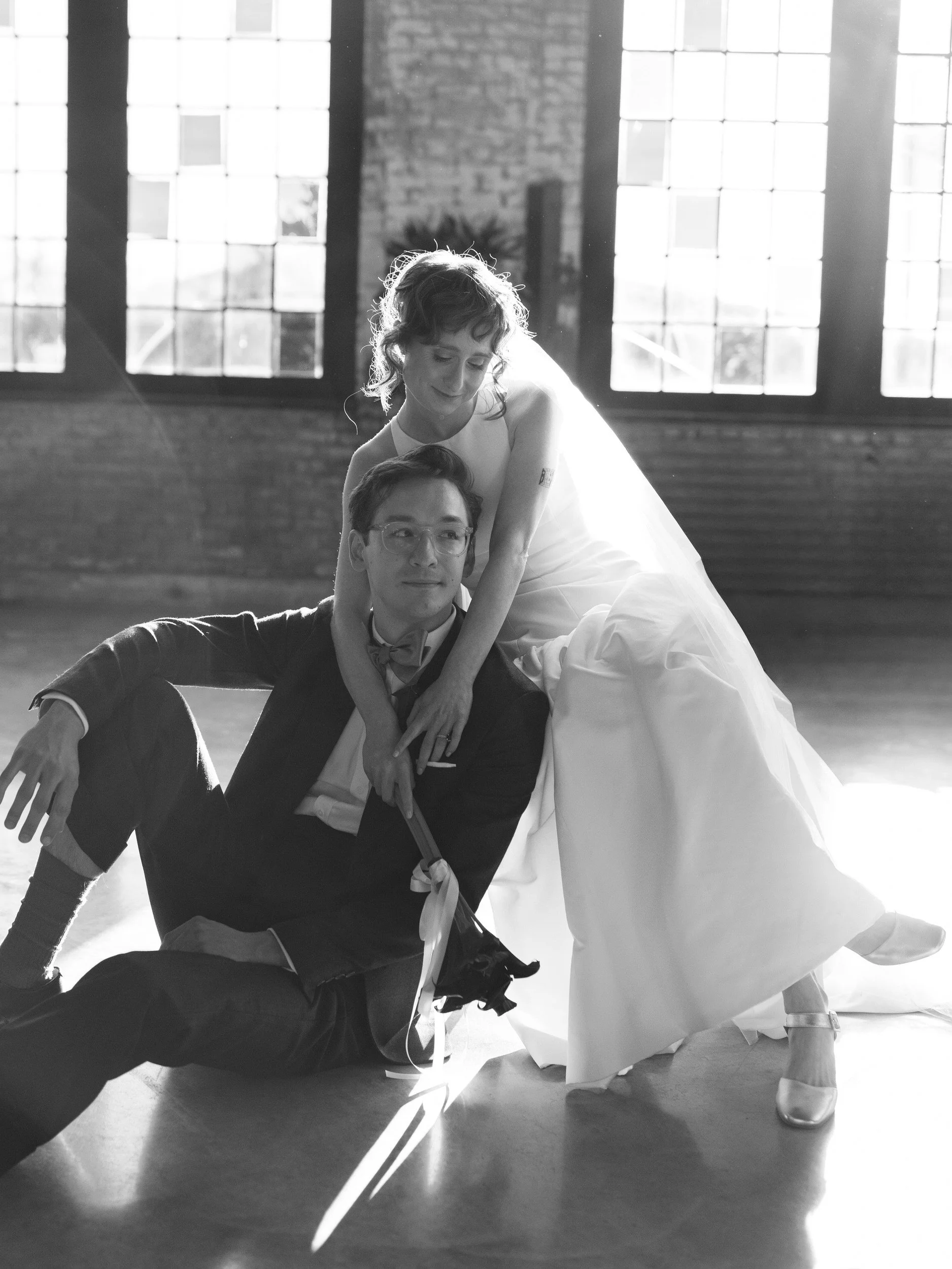 A newlywed sitting on the ground as their partner sits next to them with their arms over their shoulders 