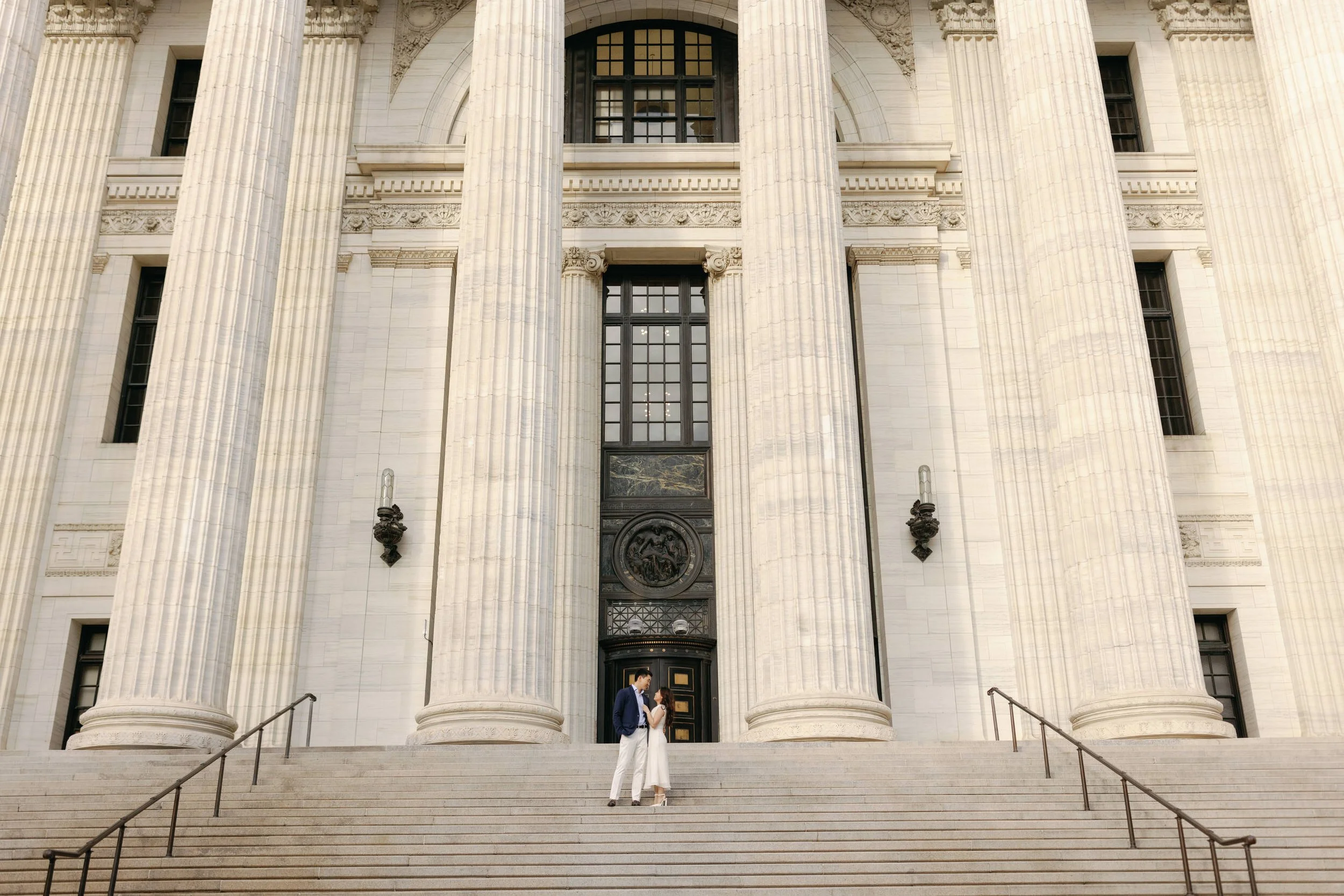 A couple smiling at each other while standing on the staircase in front of a grand building
