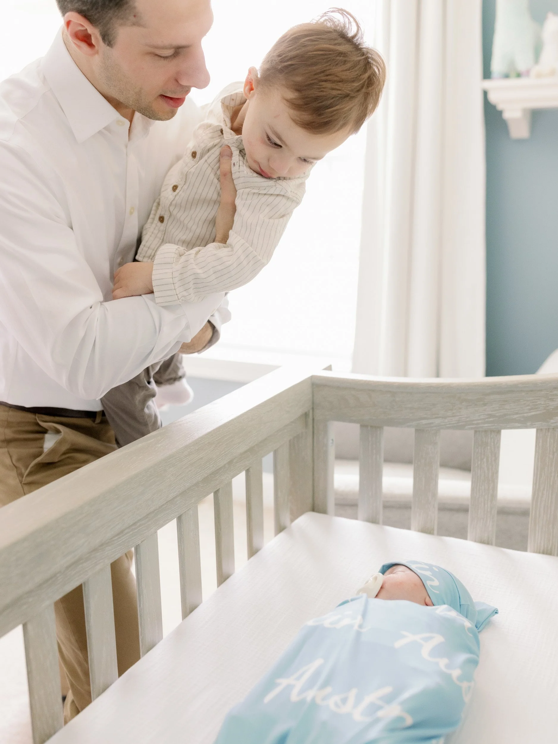 A parent holding a toddler as the look down at a newborn in a crib 