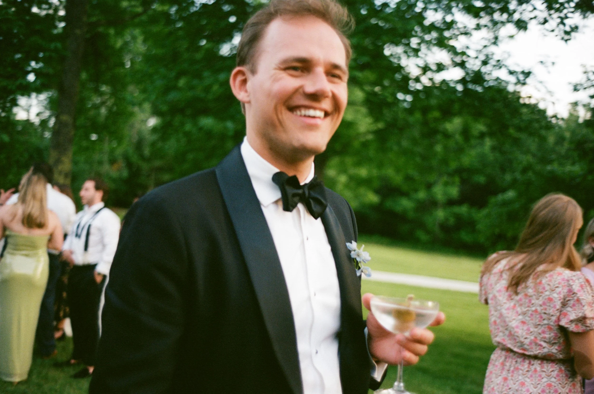a person in a suit holding a cocktail and smiling at a wedding reception 