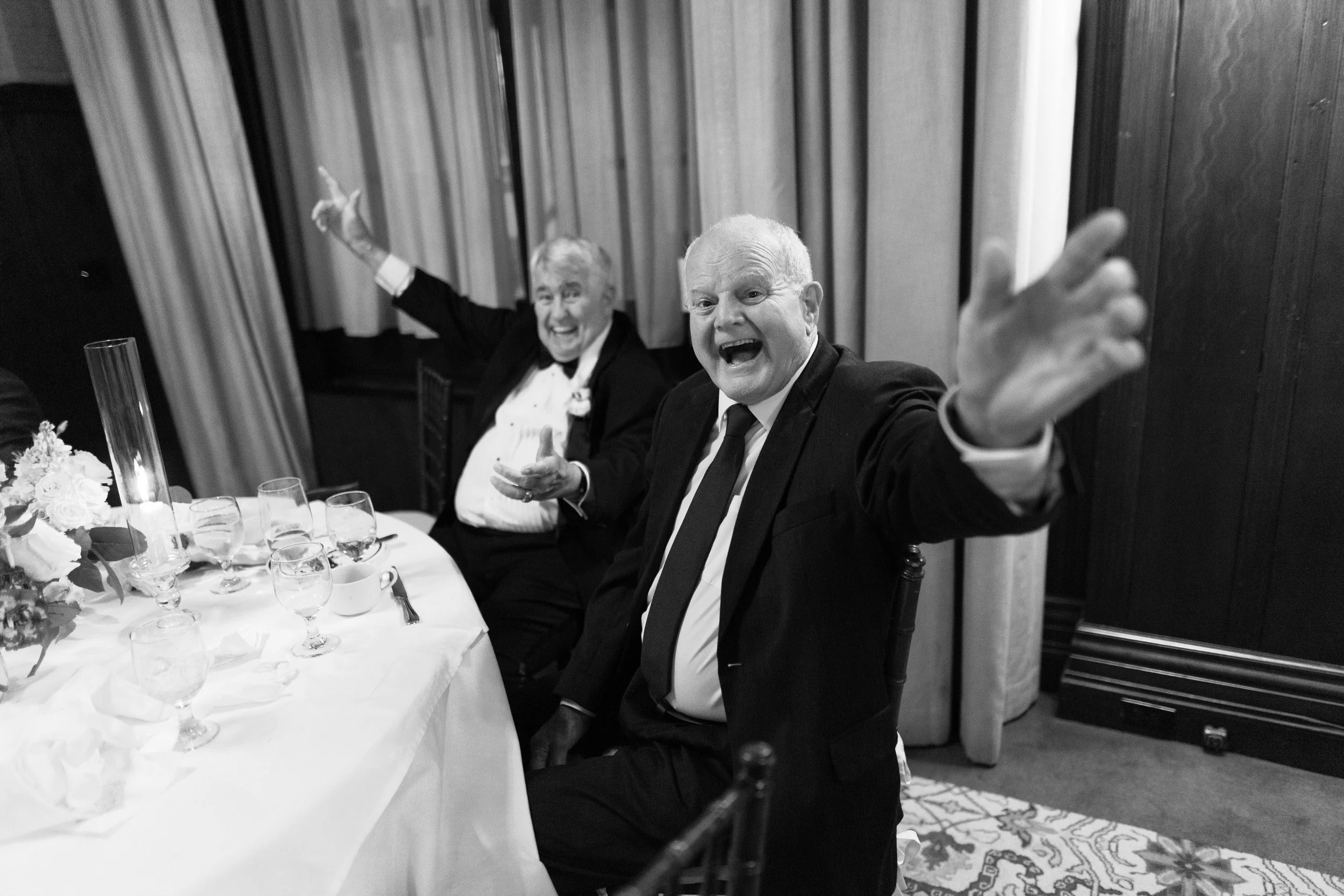 Two people in suits sitting at a table smiling with their arms outstretched