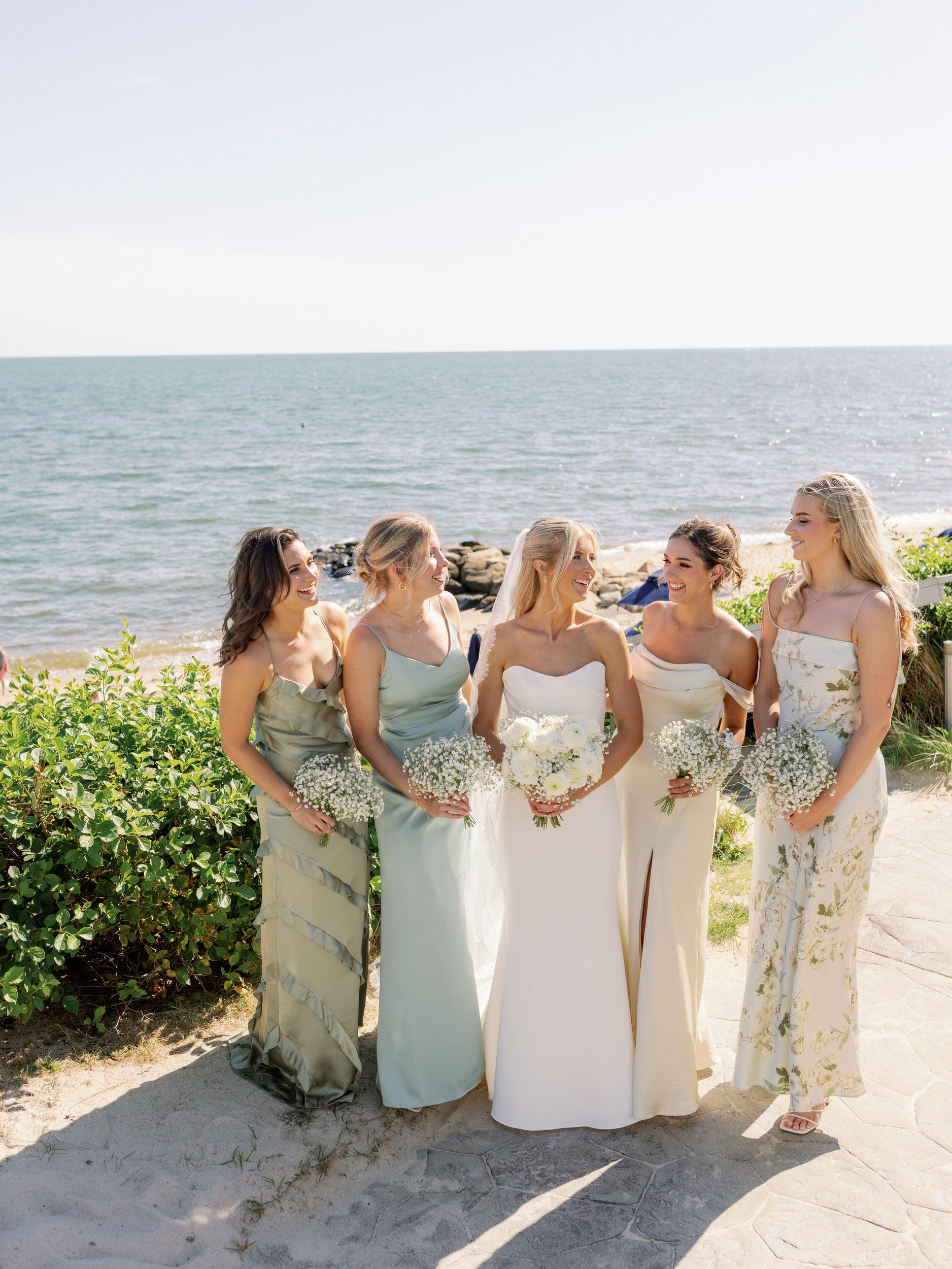 A newlywed in a wedding dress standing by the water smiling with their wedding party 
