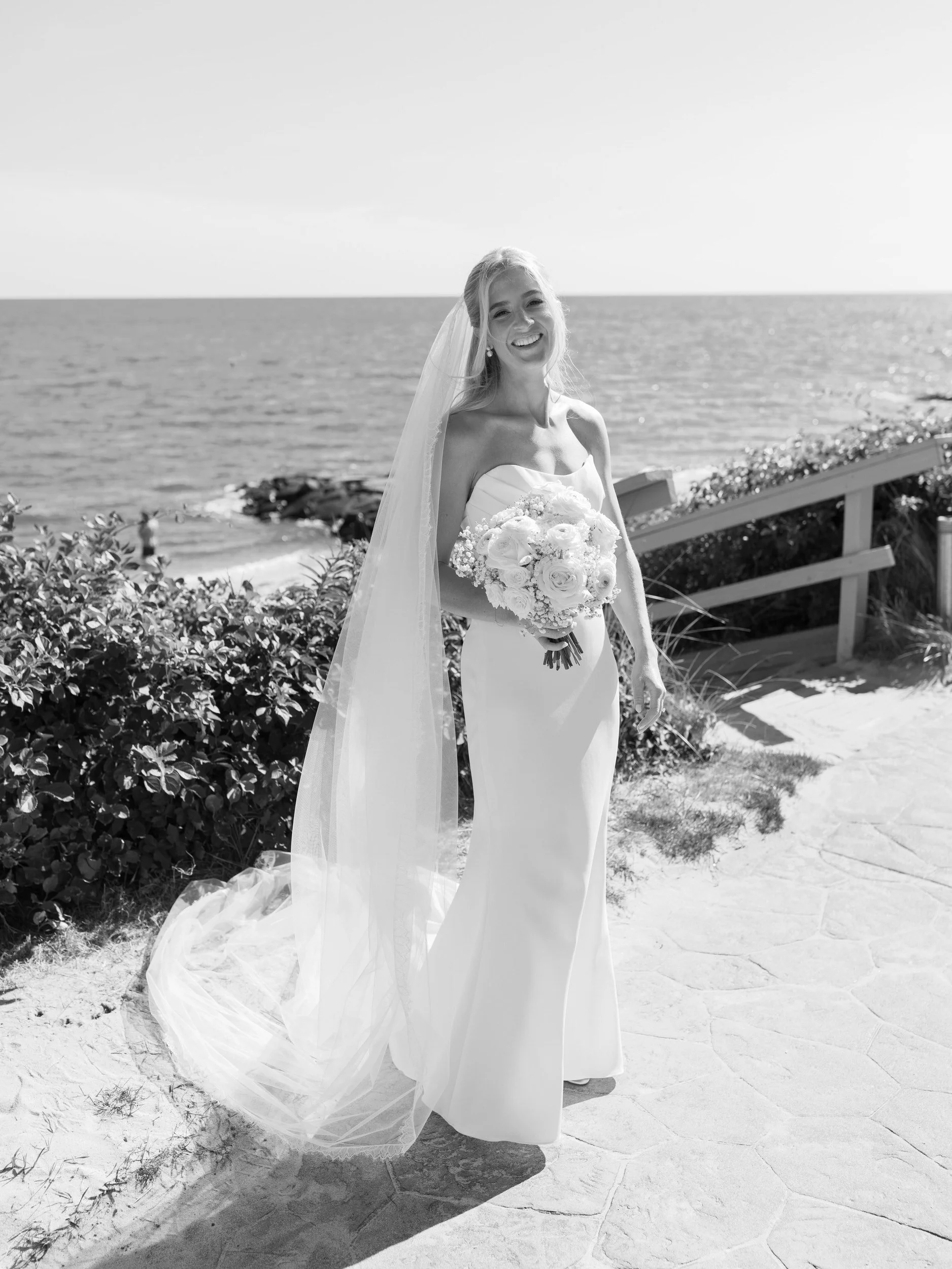 A newlywed in a wedding dress holding a bouquet of flowers and standing by the ocean 