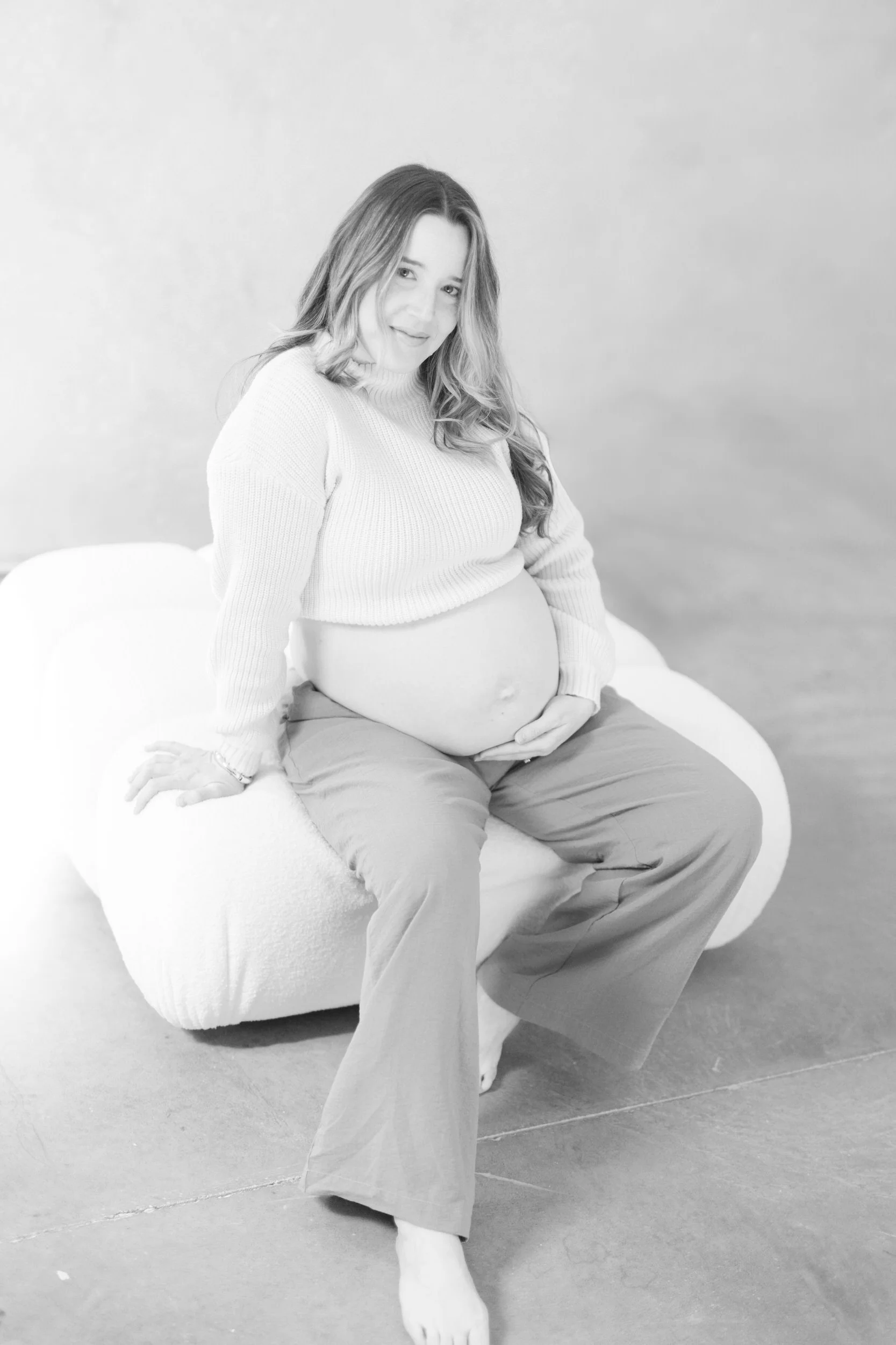 A pregnant person sitting on a small white ottoman holding their stomach 