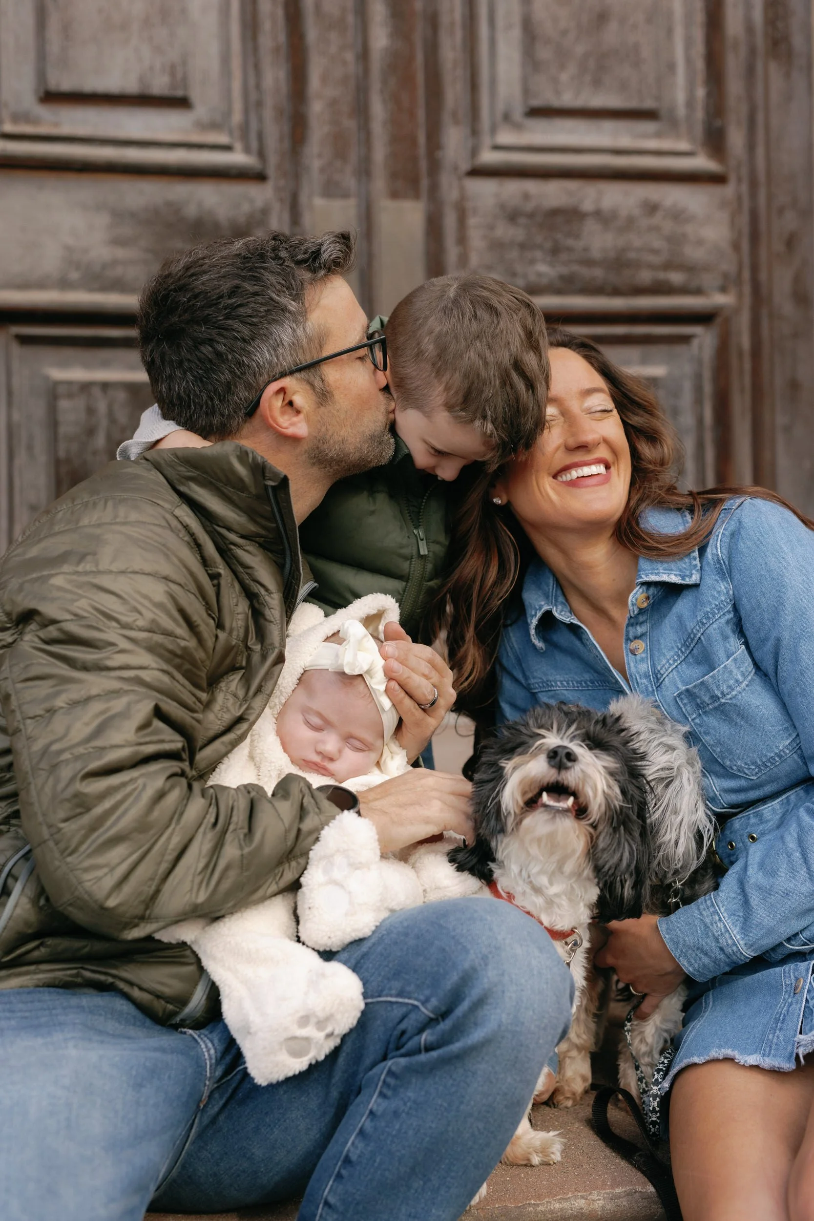 A parent holding a newborn and kissing another child's head as their partner sits on the other side of the child holding a dog