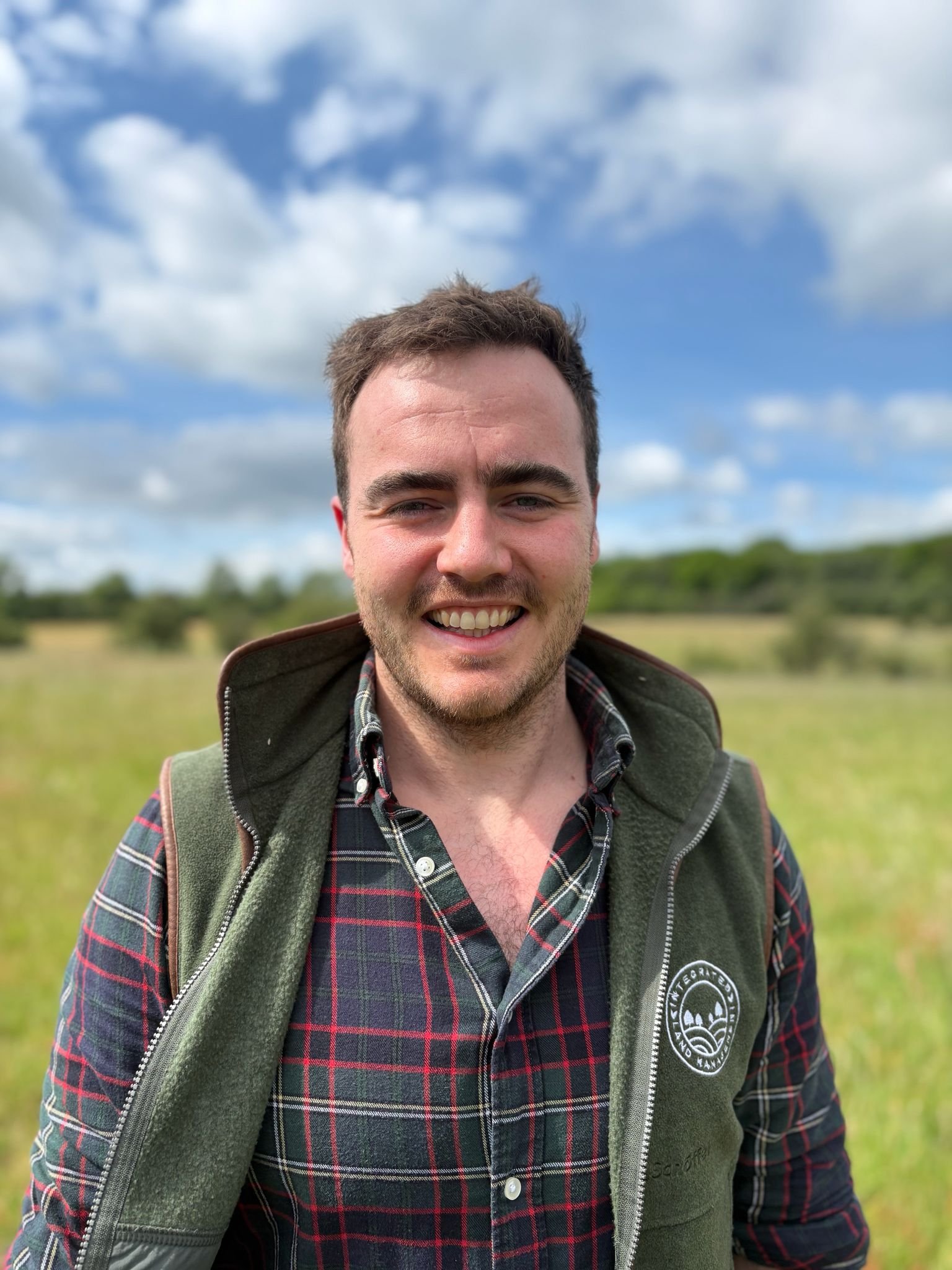 Sam smiles at the camera he has dark hair and is wearing a green ILM gilet with a countryside view in the background
