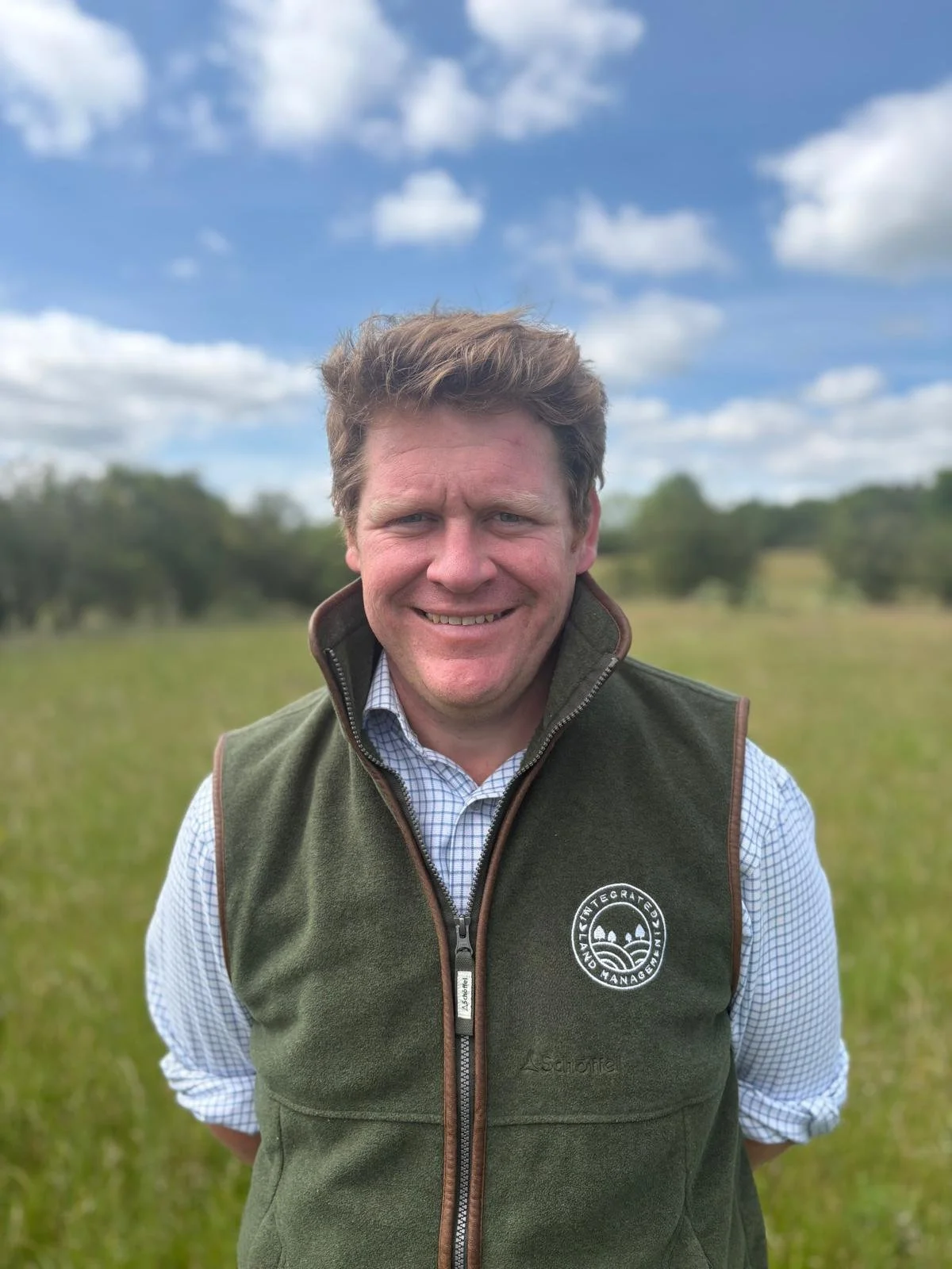 Sam is smiling to camera wearing a blue and white checkered shirt and a dark green gilet with the ILM logo on it in a field with blue skies in the background