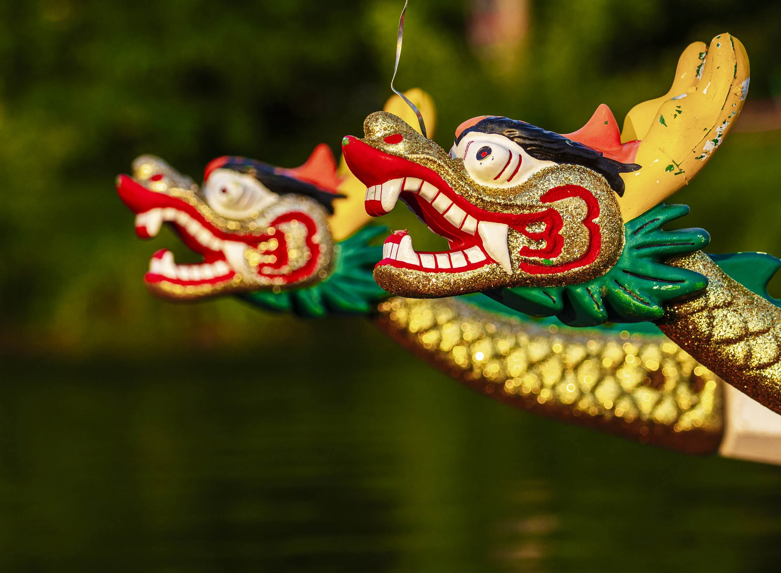 Close-up of two colorful dragon boat figurehead sculptures with gold accents, featuring open mouths, sharp teeth, and expressive eyes, mounted on a decorated boat, blurred green background.