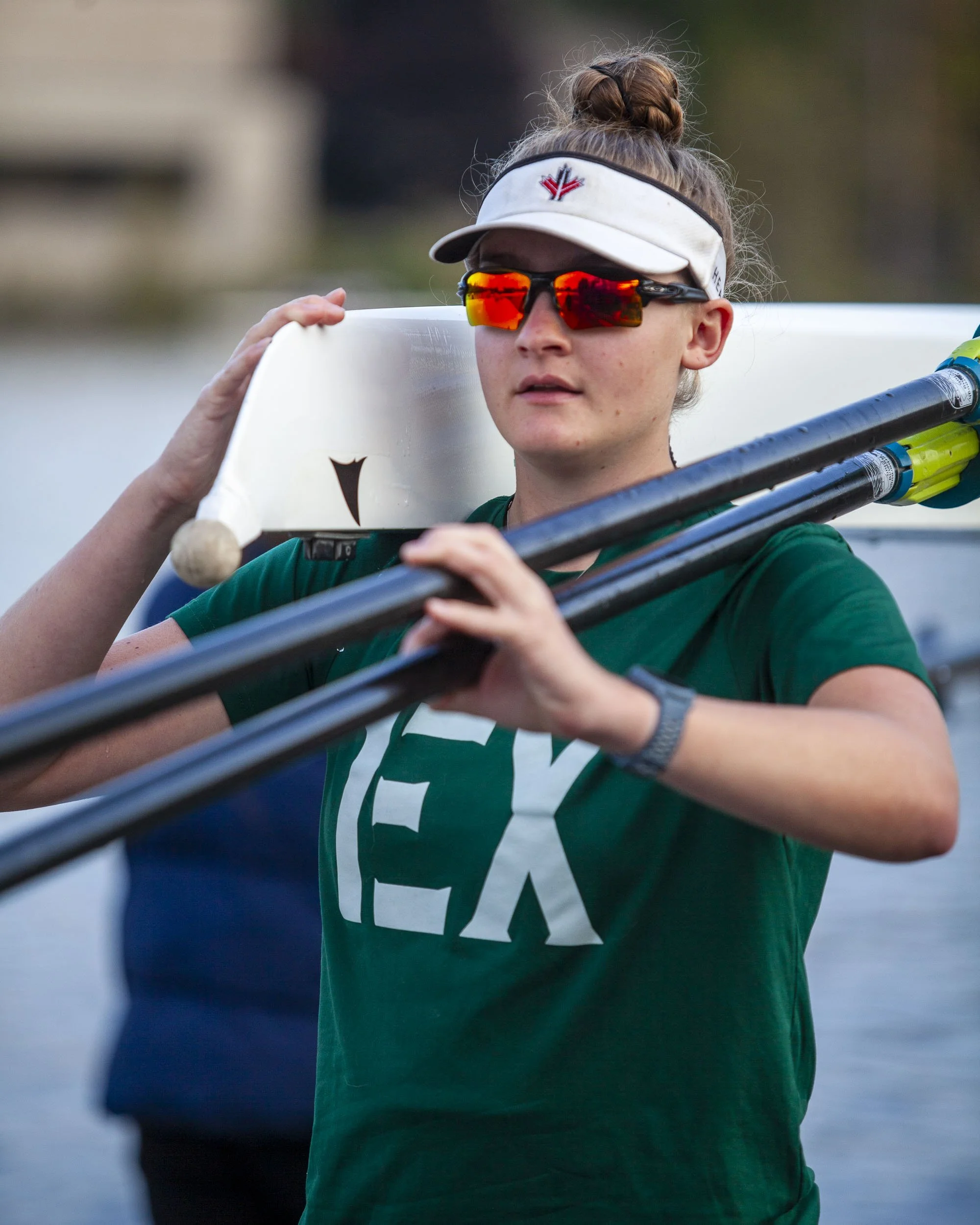 Female rower with sunglasses and a visor, carrying oars, preparing to row on a river or lake.