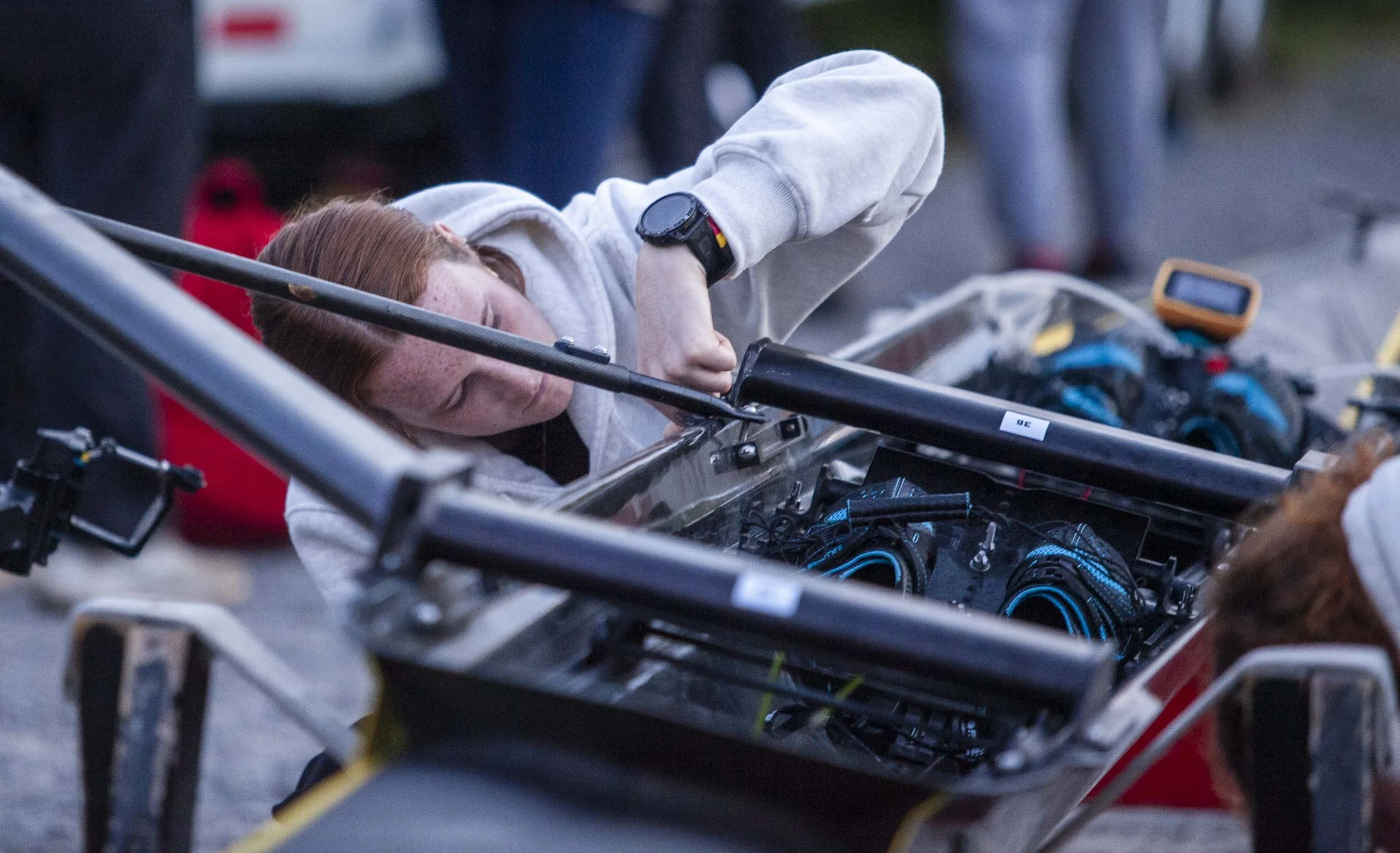 A woman with red hair working on the electronics inside a rowing shell, wearing a white sweatshirt.