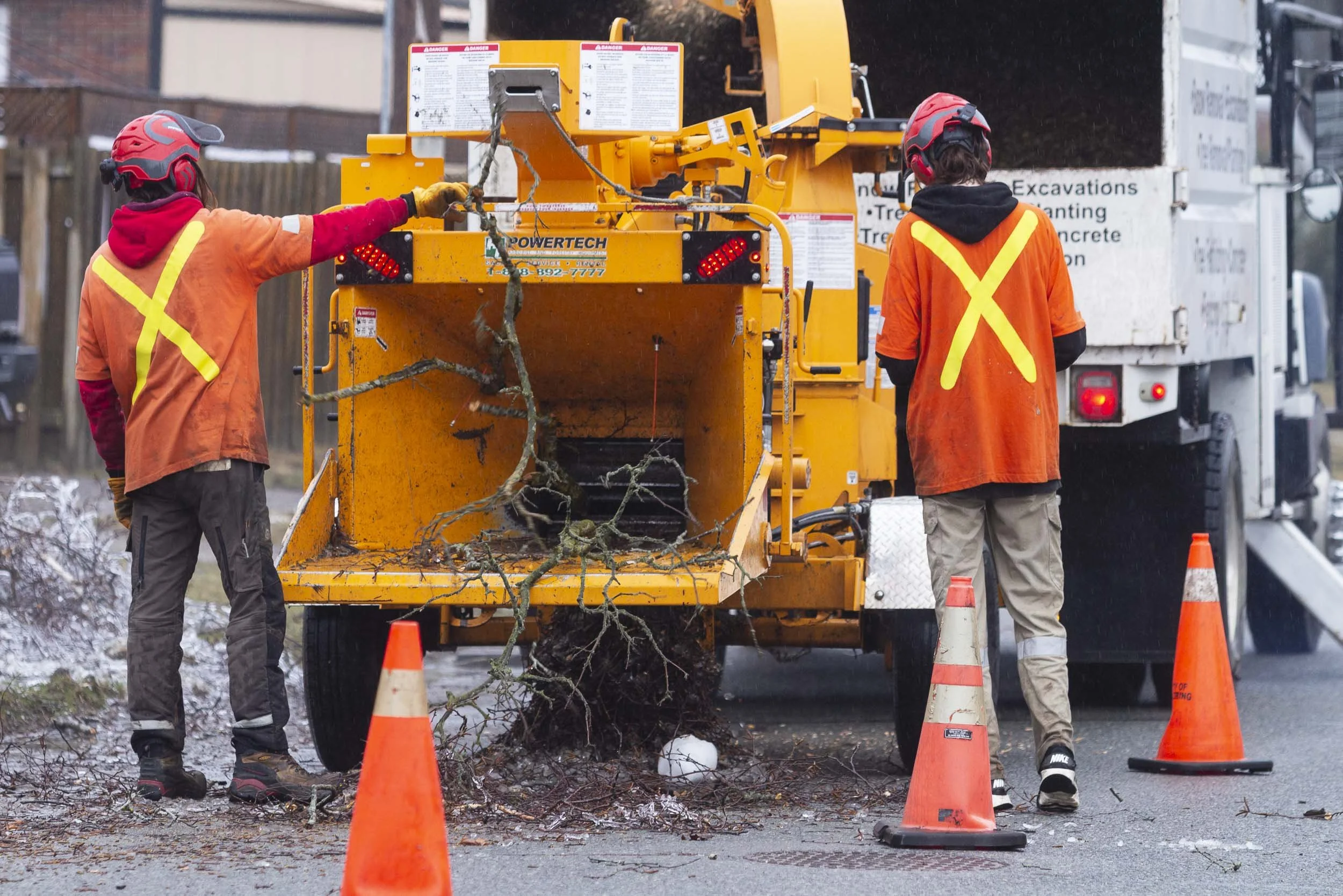 Two workers in orange safety vests and helmets operating a tree chipper on a street, removing fallen branches and debris, with traffic cones surrounding the work area.