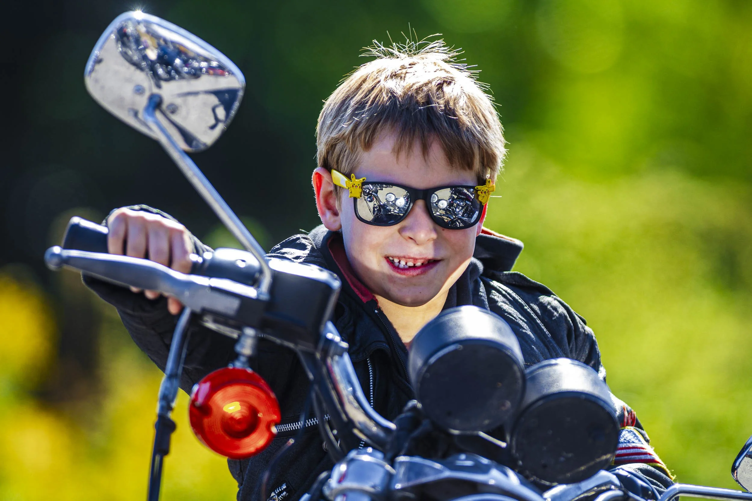 Young boy wearing sunglasses, smiling, riding a motorcycle outdoors in sunlight