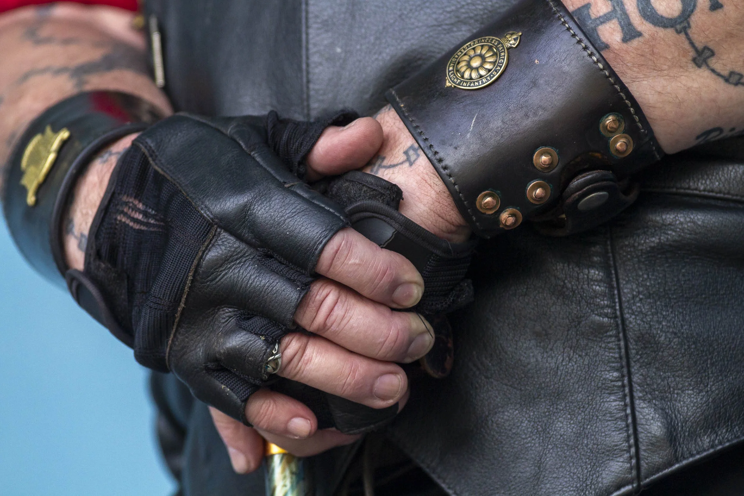 Close-up of a person's hands clasped, wearing black leather fingerless gloves, a navy leather jacket, and a black leather wristband with metal studs, revealing arm tattoos.