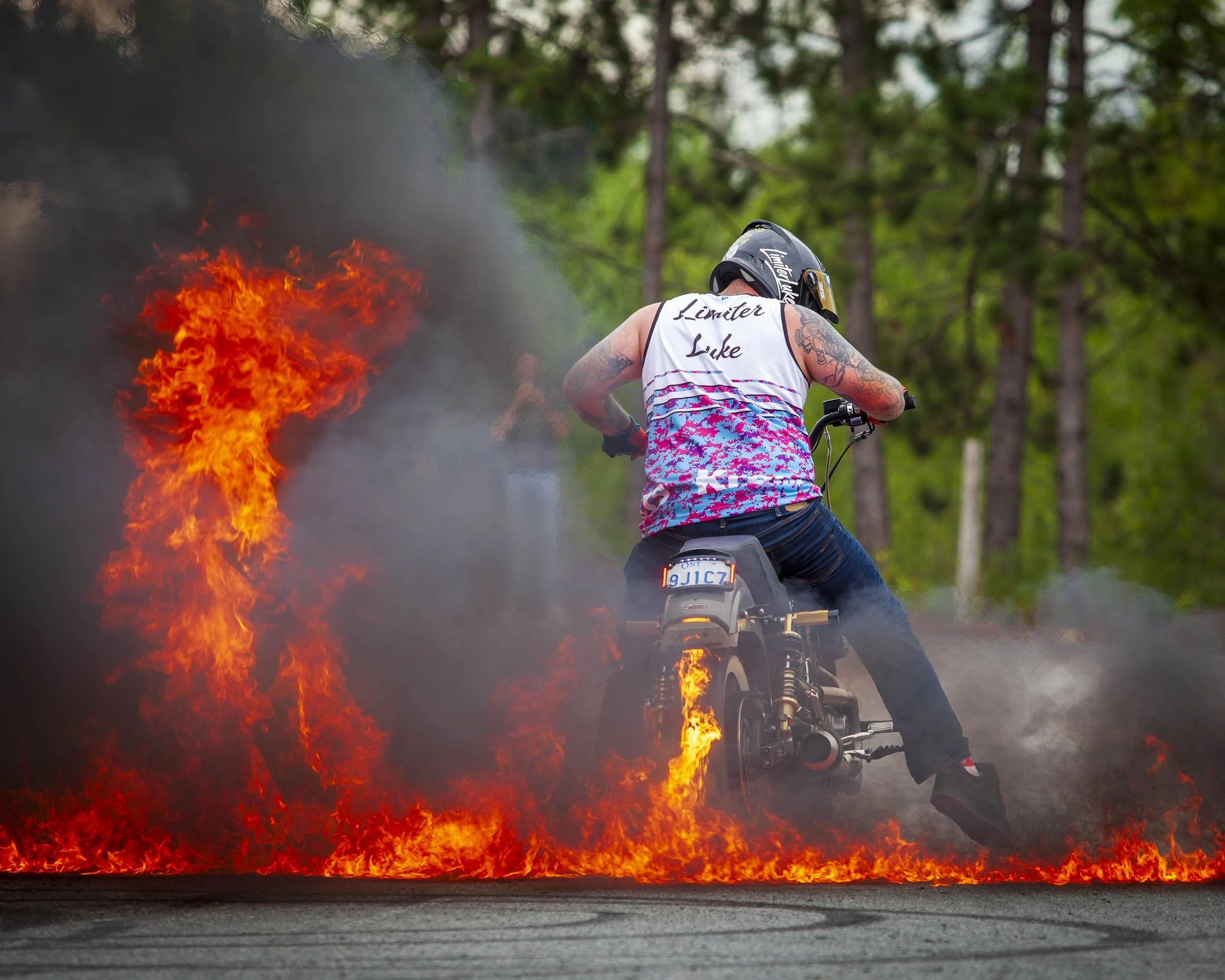 A person riding a motorcycle through a fire and smoke on a wooded road during daytime.