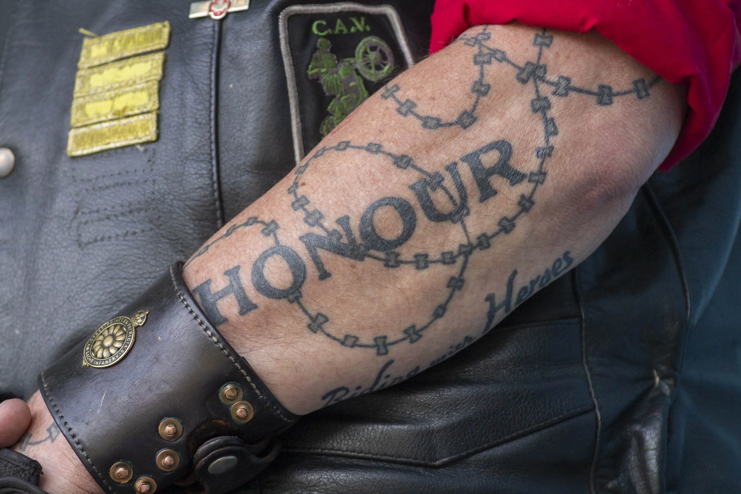 A person's tattooed arm displaying a tattoo of a heart made of chain links with the word 'HONOUR' inside, and other text including 'Heroes' and 'Respect' partially visible. The person is wearing a black leather wristband and a leather vest with patch