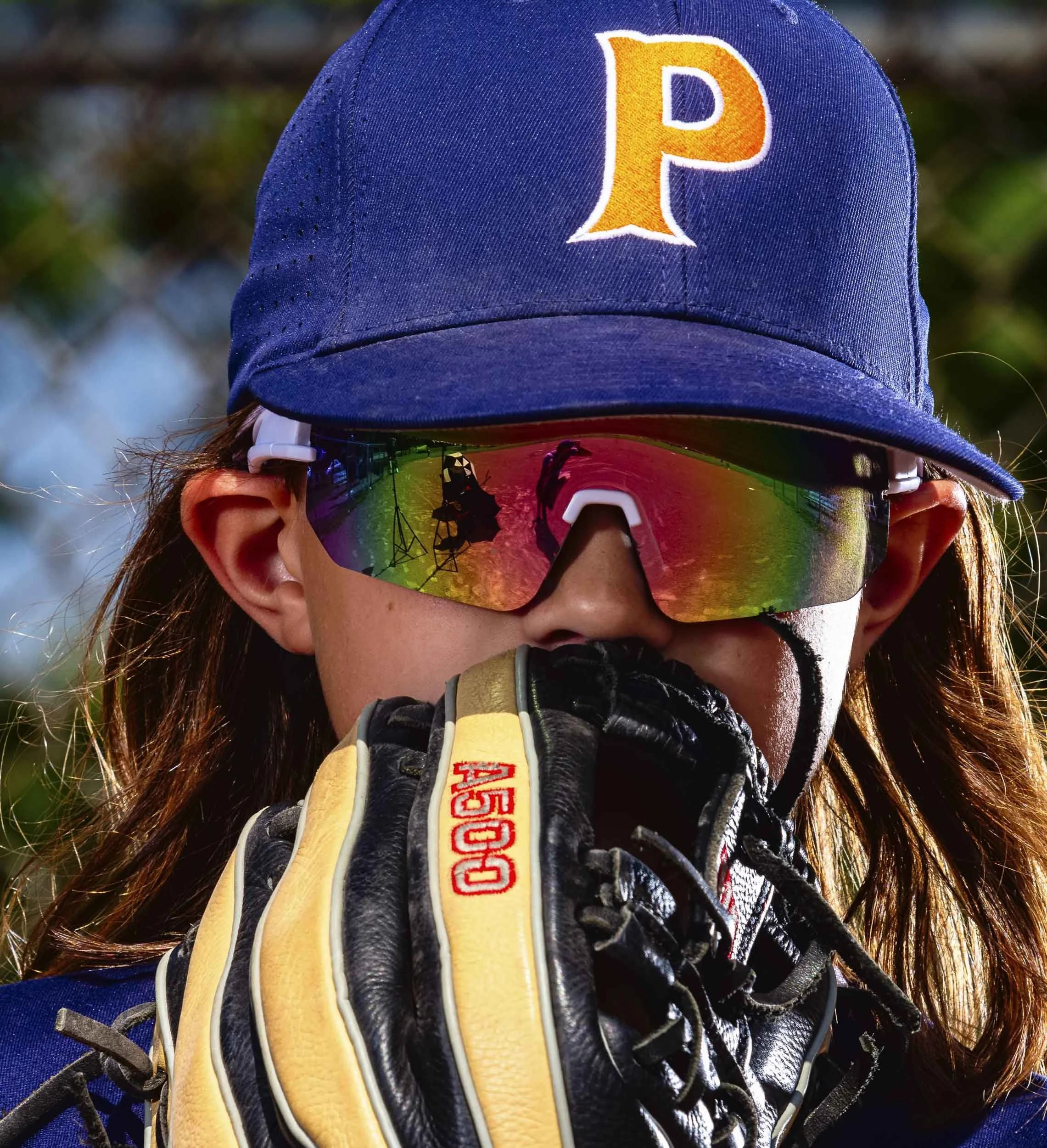 Close-up of a boy wearing a blue baseball cap with a yellow letter P, reflective sports sunglasses, black and yellow baseball glove, and a black face mask, outdoors.