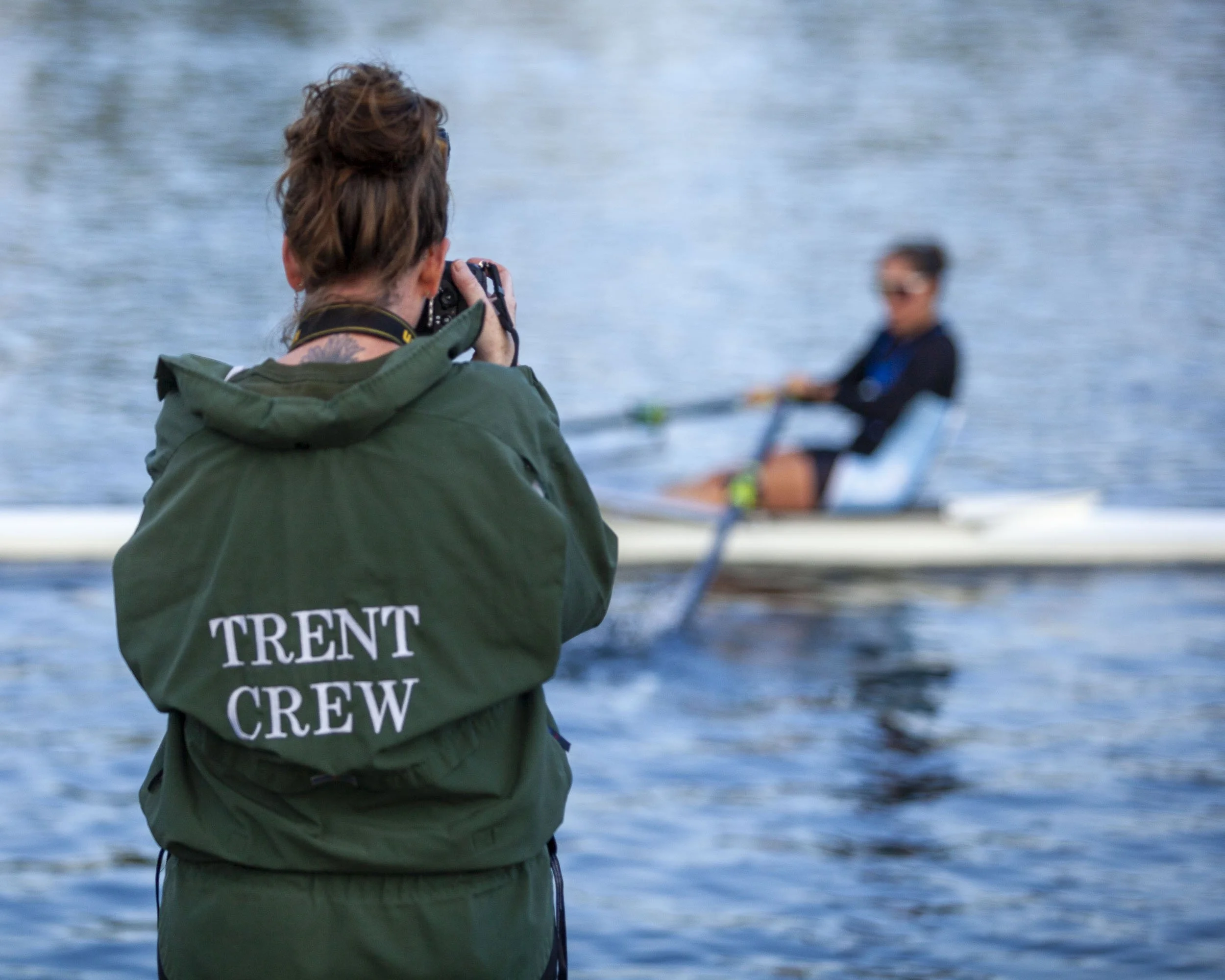A person in a green jacket labeled "Trent Crew" taking a photo of a rower in a boat on a body of water.
