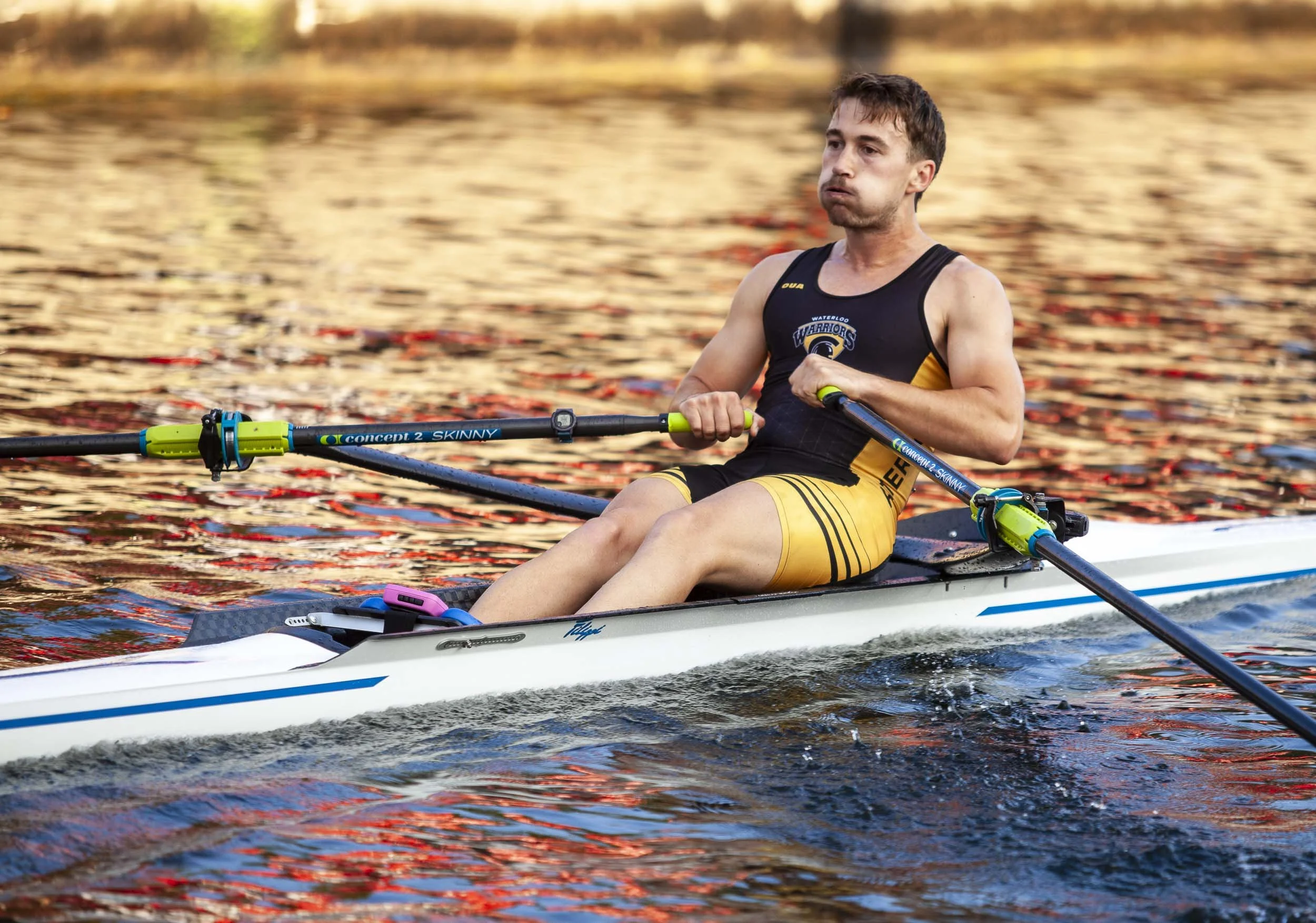 A man in a yellow and black rowing uniform paddles a racing shell with oars on a calm lake during sunset.