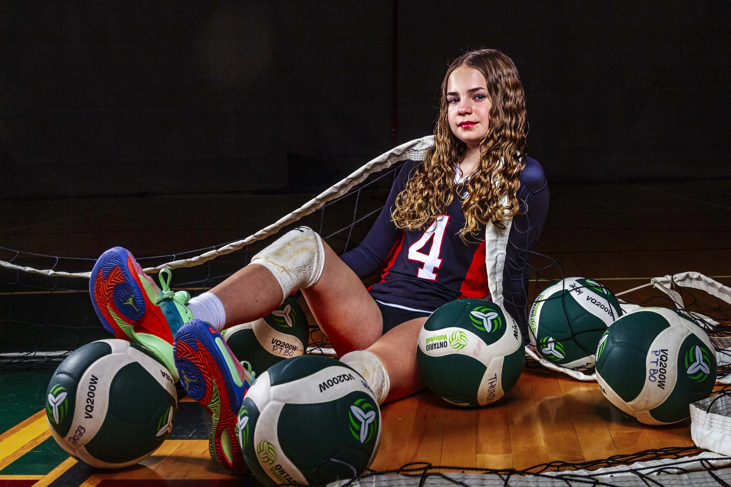 Young female volleyball player sitting on gym floor surrounded by volleyballs, wearing a navy blue and red sports uniform with the number 14, athletic shoes, and knee pads, with a volleyball net behind her.