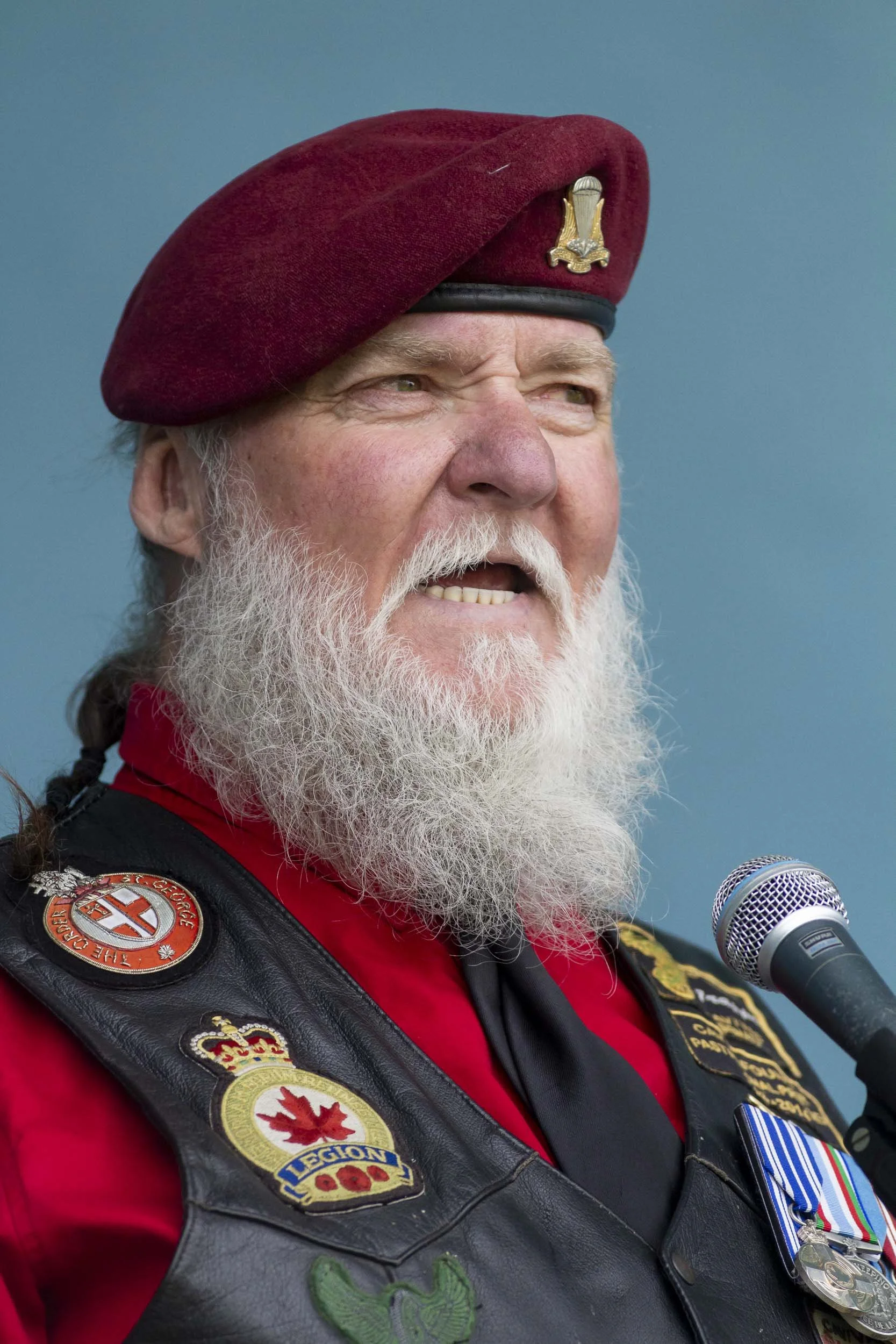 An older man in a red beret and leather vest decorated with patches and medals, speaking into a microphone against a blue background.