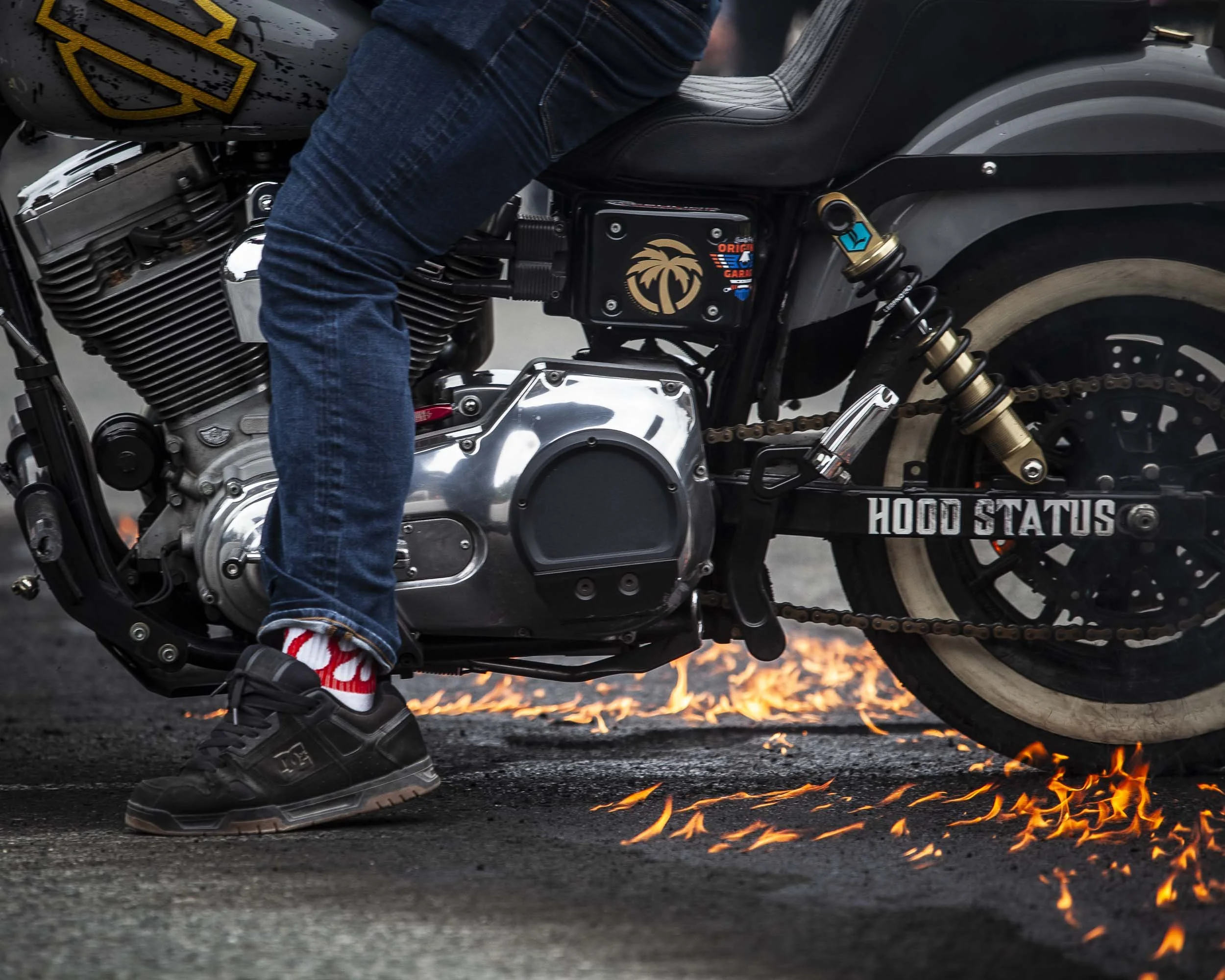 A person riding a black motorcycle with chrome details, wearing jeans and black sneakers with red and white socks, on a road with sparks coming from underneath the bike.