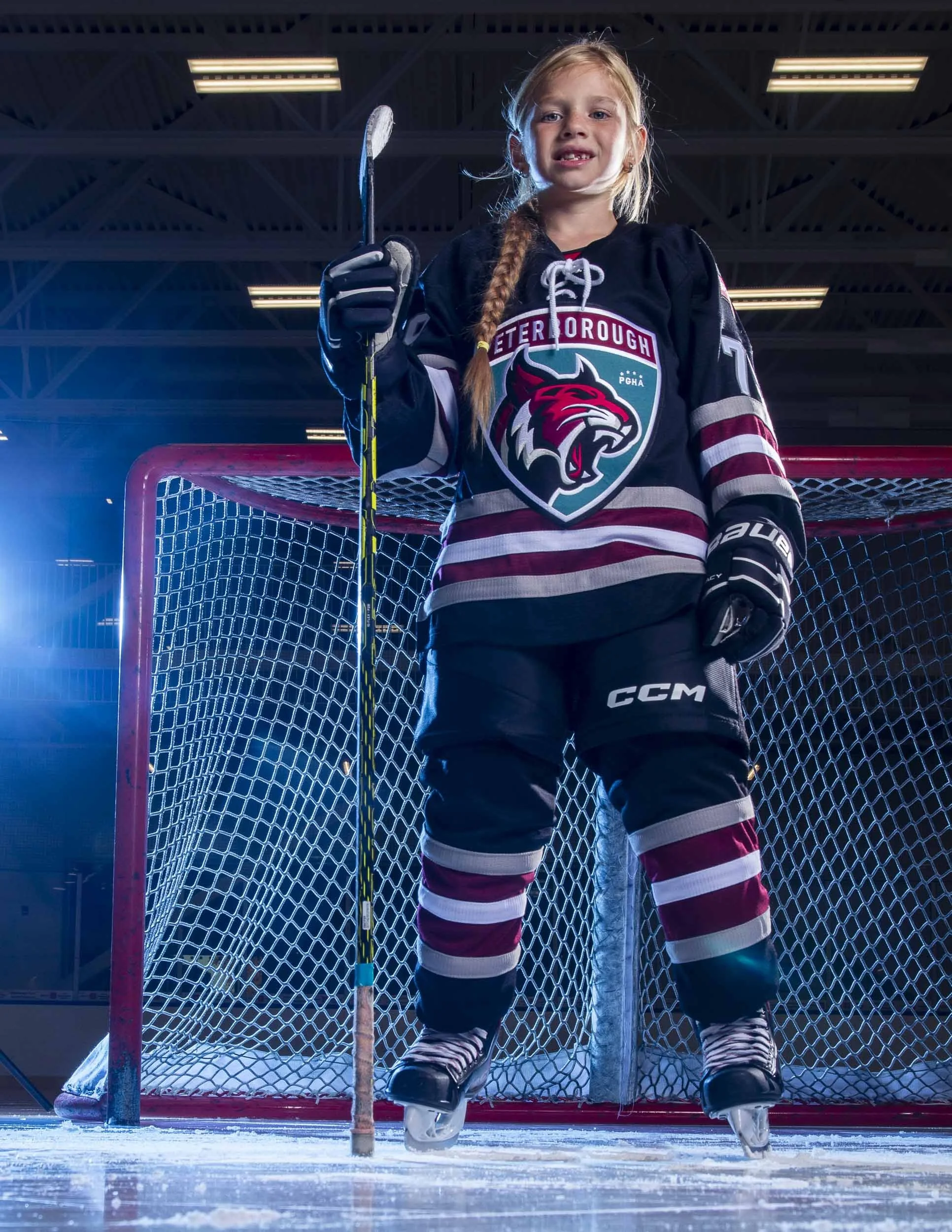 Young girl in hockey gear standing on ice rink in front of goal, holding hockey stick, wearing a Peterborough Petes jersey and skates.