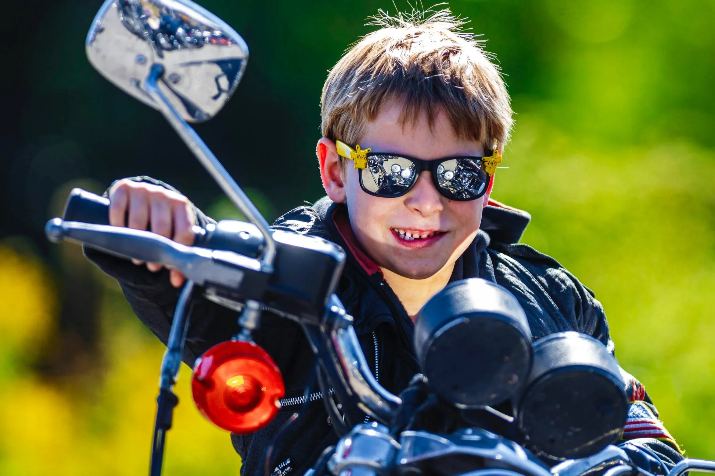 Young boy wearing sunglasses and a black jacket, riding a motorcycle outdoors on a sunny day. The background is blurred with green foliage.