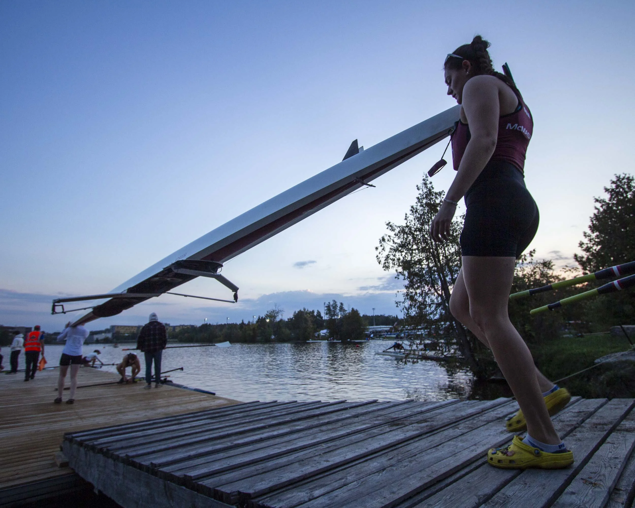 A woman in a maroon tank top and black shorts is walking across a wooden dock toward a large racing shell or boat at twilight. Several other people are on the dock, preparing for rowing by the water.