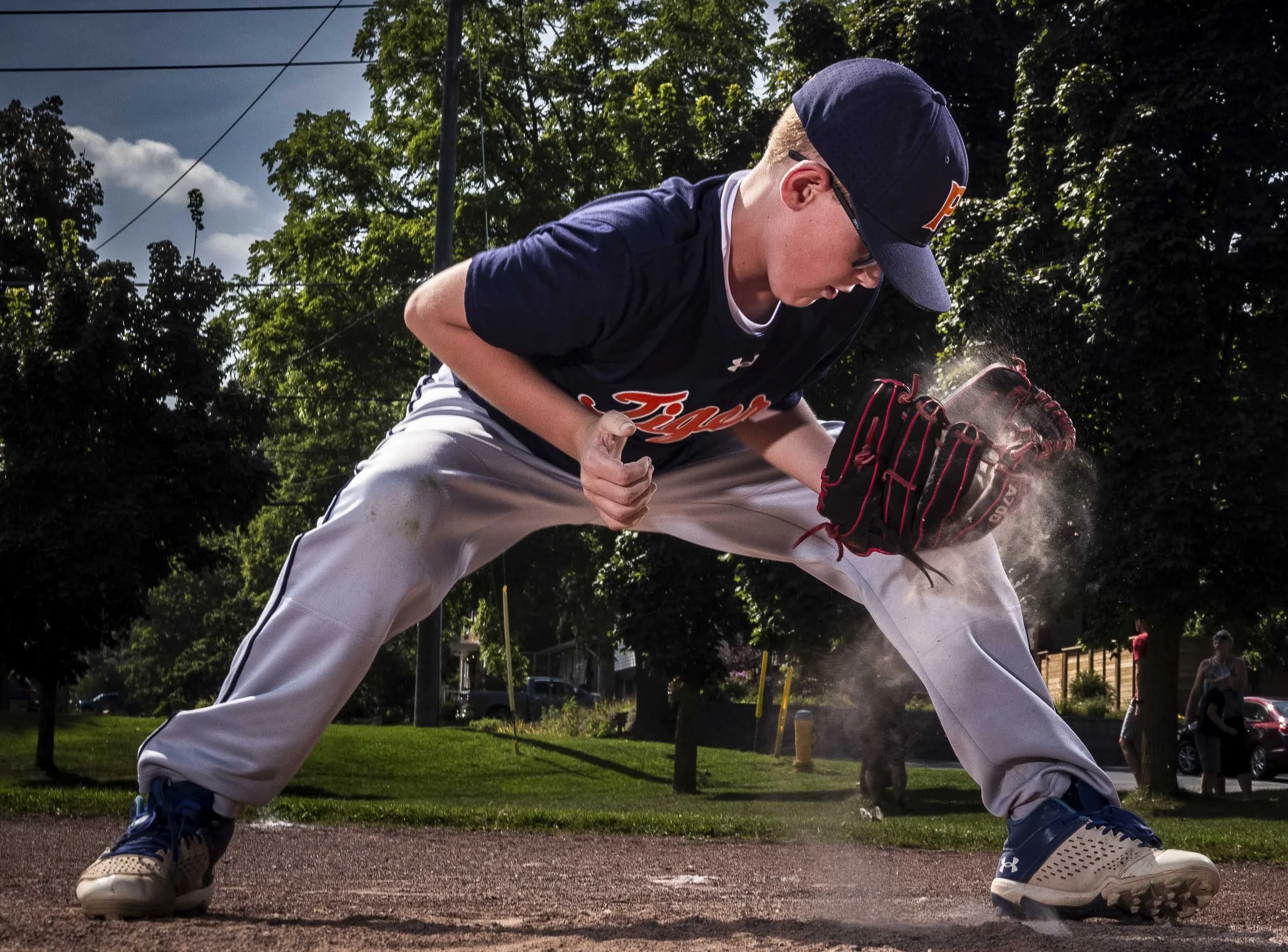 A young baseball player wearing a navy blue jersey, gray pants, and a cap, fielding a ground ball on a dusty baseball field with trees and a utility pole in the background.
