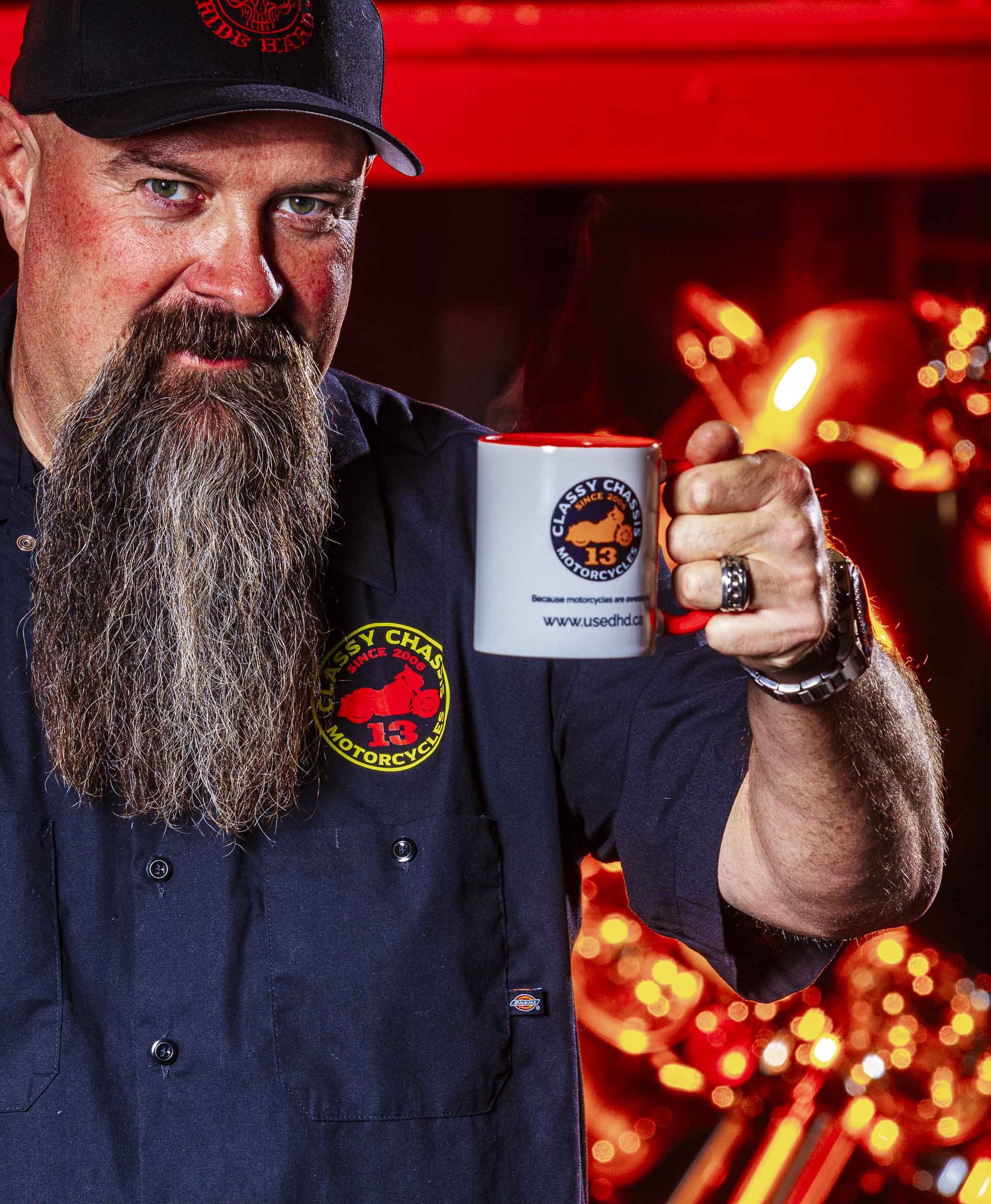 A man with a long beard and mustache wearing a black cap and dark shirt, holding a steaming white mug with a logo that reads 'Classy Chassis Motorcycle 13'. The background is warm with blurry orange and yellow lights.